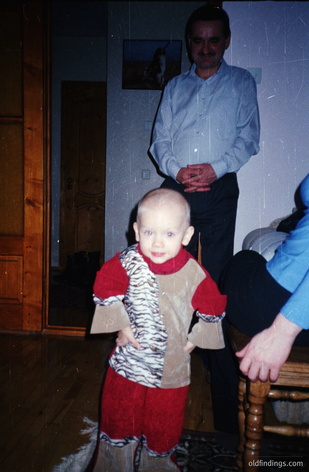 A young child, likely a toddler, stands in a warmly-lit interior, wearing a playful animal-print outfit. A man in a button-down shirt stands behind, hands clasped. Appears to be a candid, casual portrait, possibly a family snapshot. Likely 1970s or 80s given the color palette and clothing styles. Interior suggests a traditional home.