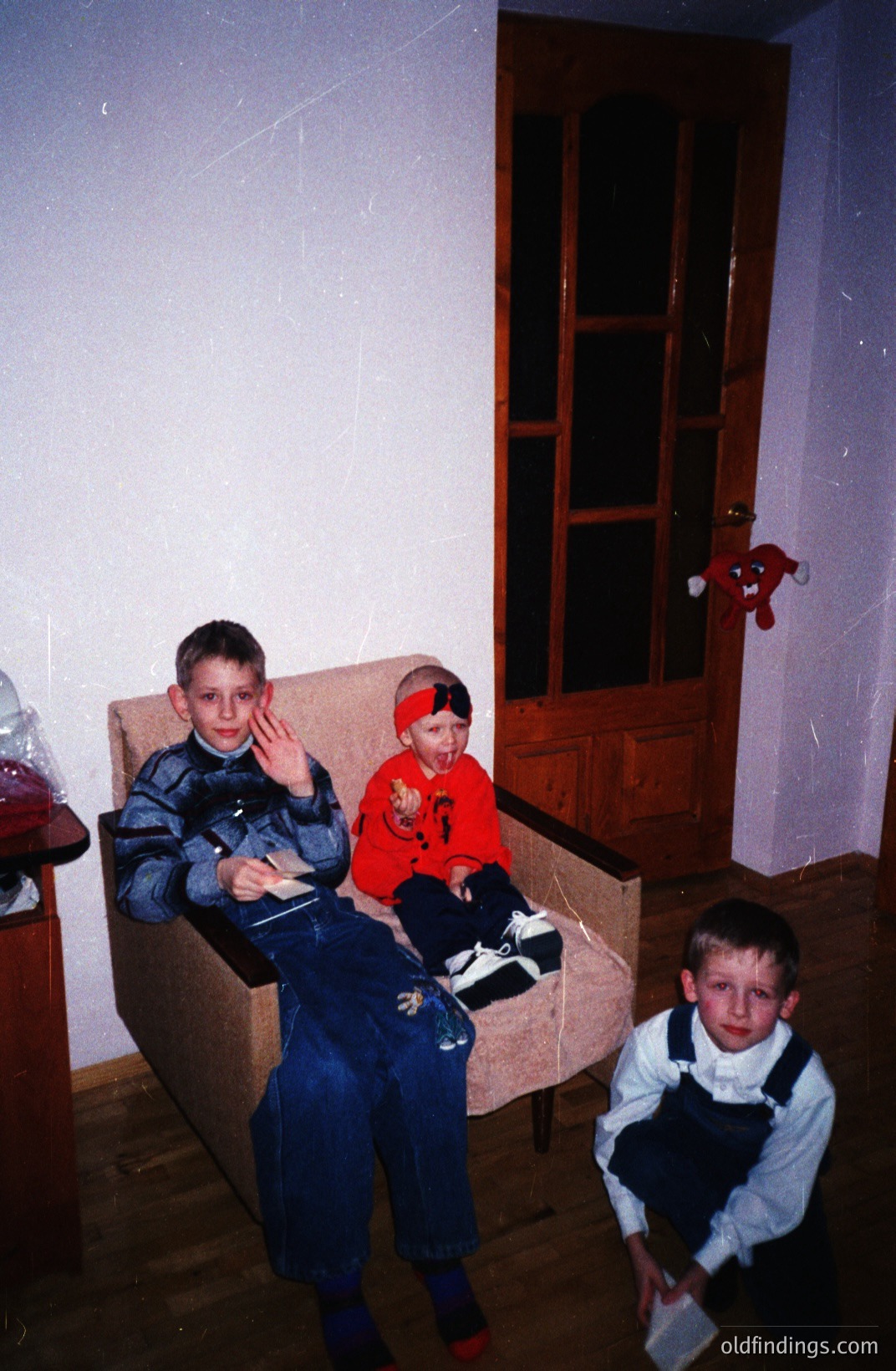 Three boys posed in a room with a patterned armchair, paneled door, and visible dust particles in the air. Two boys are seated; one waving, the other bundled in a red sweatshirt. A third boy stands, holding what appears to be a tissue. Likely taken in the 1980s or 90s, based on clothing styles. Appears to be a candid family snapshot.