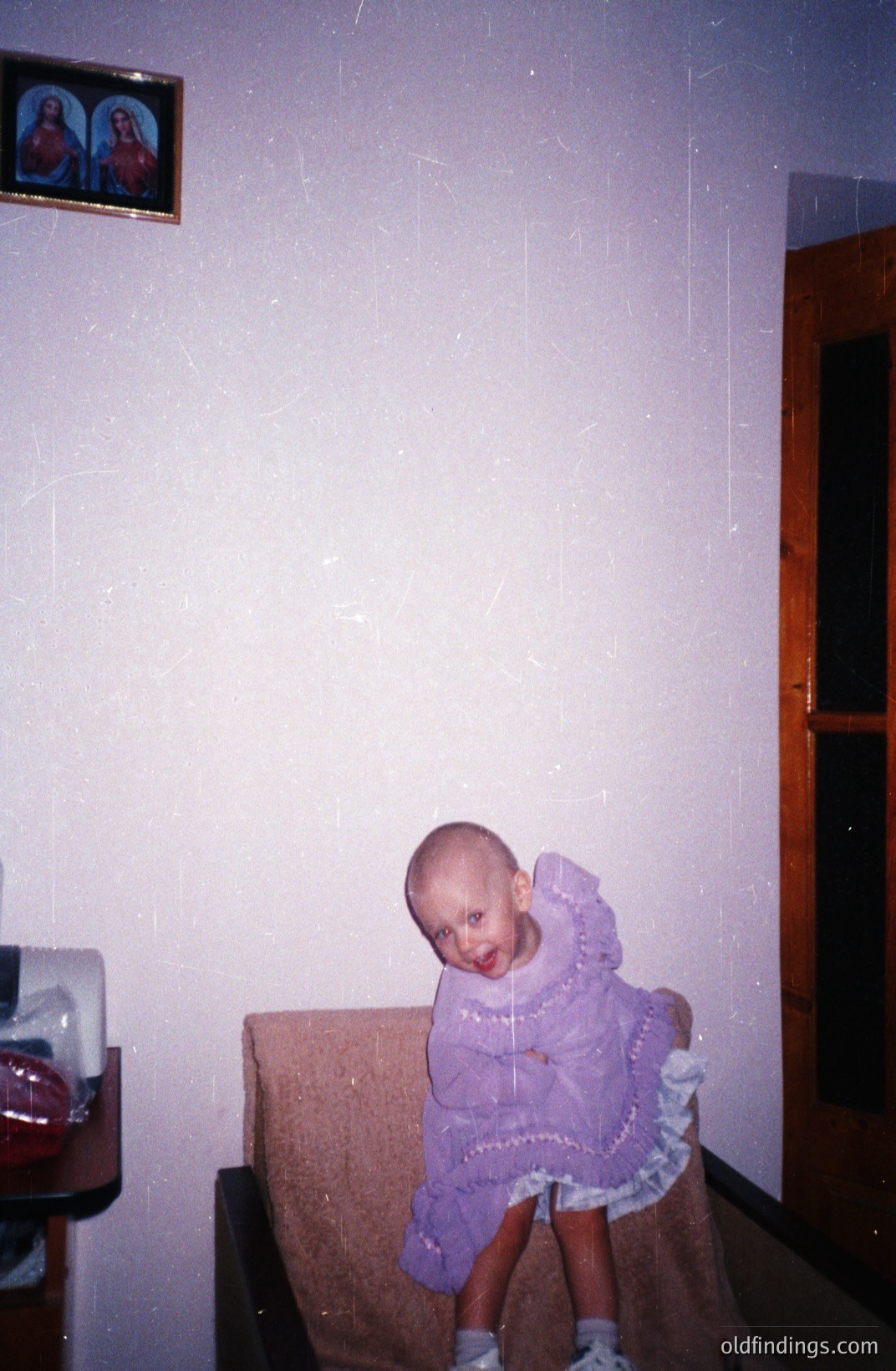 A young child, likely a toddler, seated on a cushioned chair wearing a lavender dress. The room features a stark white wall adorned with a framed religious icon. The style suggests a domestic interior, potentially Eastern European. Likely a candid snapshot from the 1970s or 1980s.