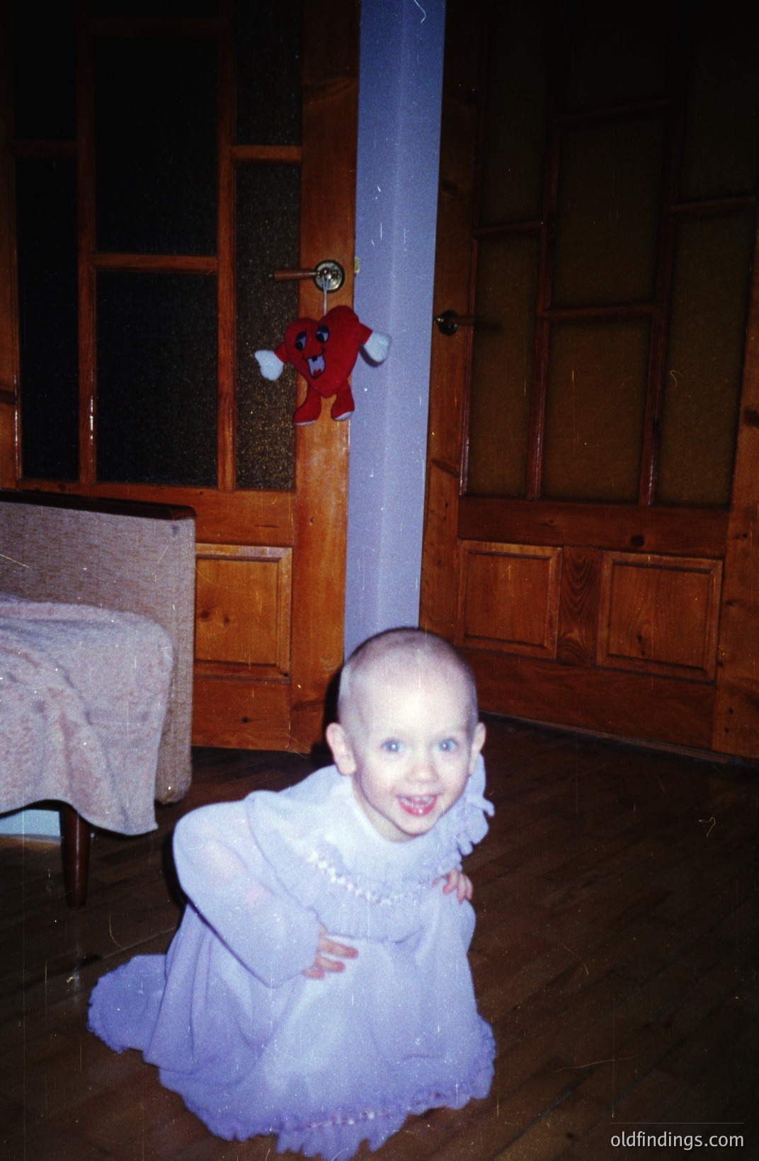 A young child with a shaved head kneels in a warmly-lit room, wearing a light purple, ruffled dress. A plush toy hangs on a door beside them. Likely a family snapshot, capturing a moment of childhood. Estimated 1970s, based on color palette & aesthetic.