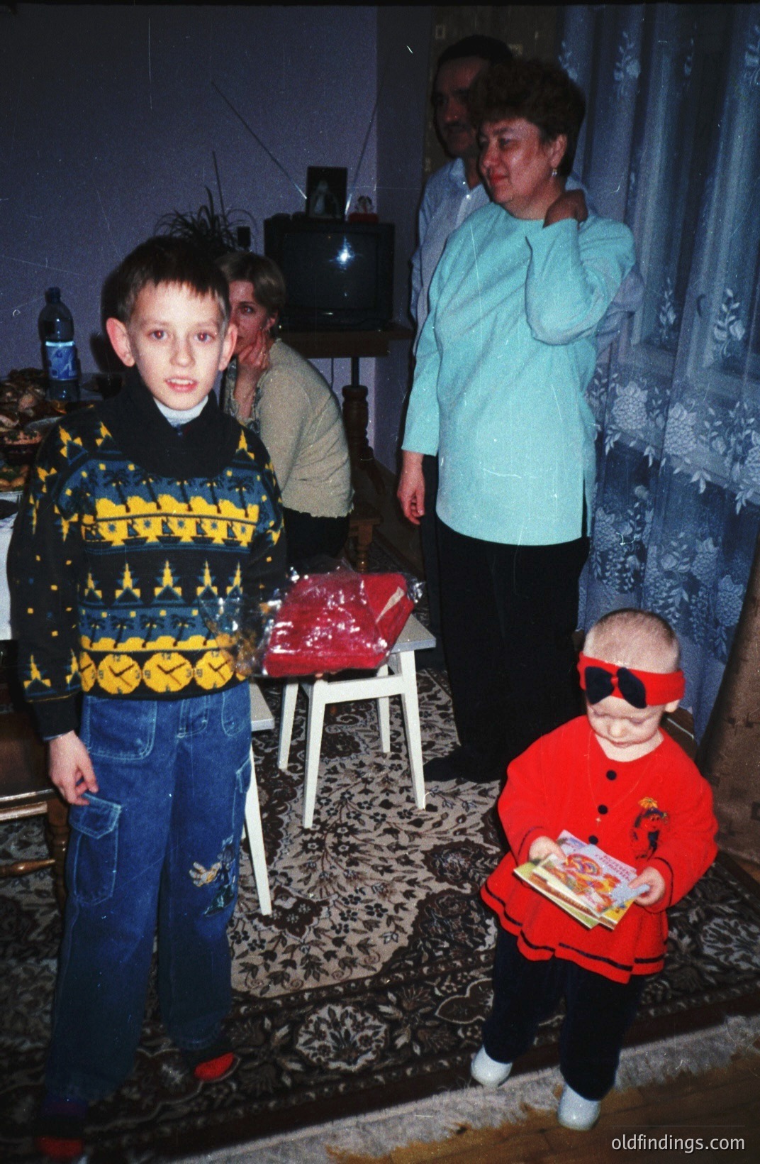 A young boy in a cable-knit sweater and jeans stands next to a toddler in a red tracksuit and sunglasses, both holding snacks. A woman with a large perm stands behind, appearing to supervise. Interior scene with a television and patterned rug. Likely 1980s or early 1990s.