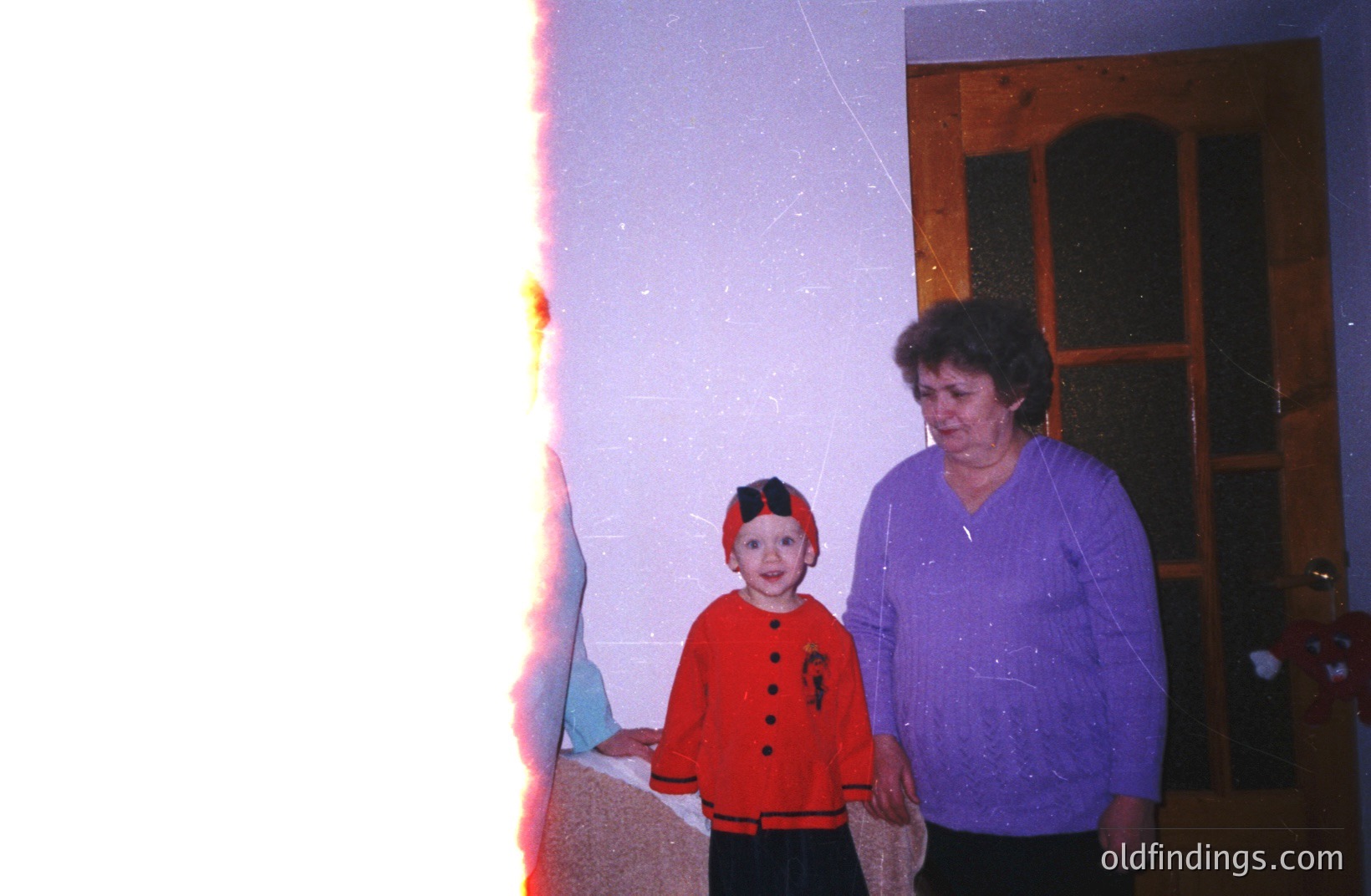 A young child in a red and black costume stands alongside an adult woman within a doorway. The photograph’s dated appearance suggests a snapshot from the 1970s or 1980s. Interior architecture shows dark wood paneling. Likely a family portrait. Potential for historical research on childhood fashion.