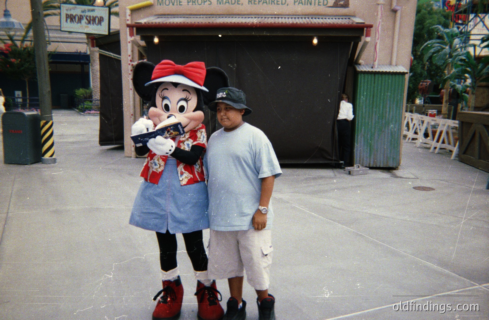 A young boy stands next to a costumed character resembling Minnie Mouse, posing for a photo. The backdrop suggests a themed area, possibly a film studio or amusement park, featuring vintage signage. Likely taken in the 1990s, the attire—boy’s graphic tee and bucket hat—reflects that era. A valuable nostalgic image for merchandise or marketing.