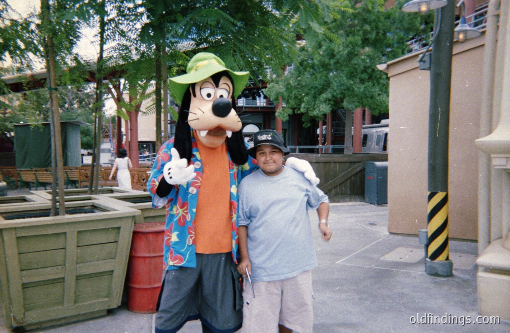 A man poses with Goofy, the Disney mascot, in an outdoor park setting, likely Disneyland or Disney World. Goofy wears a Hawaiian shirt and a floppy hat. The man wears a baseball cap and casual attire. Lush greenery and buildings are visible in the blurred background. Likely taken in the 1990s or early 2000s.