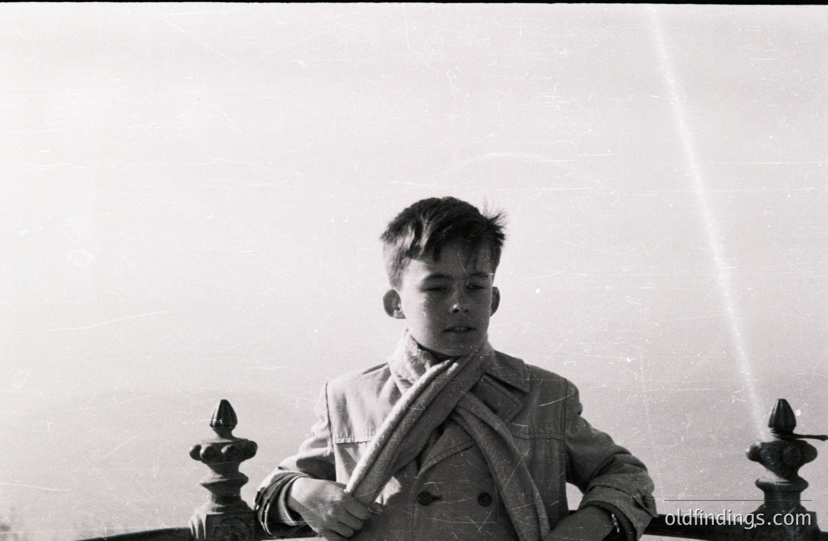 A young boy stands behind an ornate wrought-iron railing, gazing downward. He wears a double-breasted coat and scarf. The image exhibits signs of age, including grain and surface scratches. Likely a candid portrait, circa 1960s, possibly in a European setting. A strong compositional element, emphasizing youth and reflection.