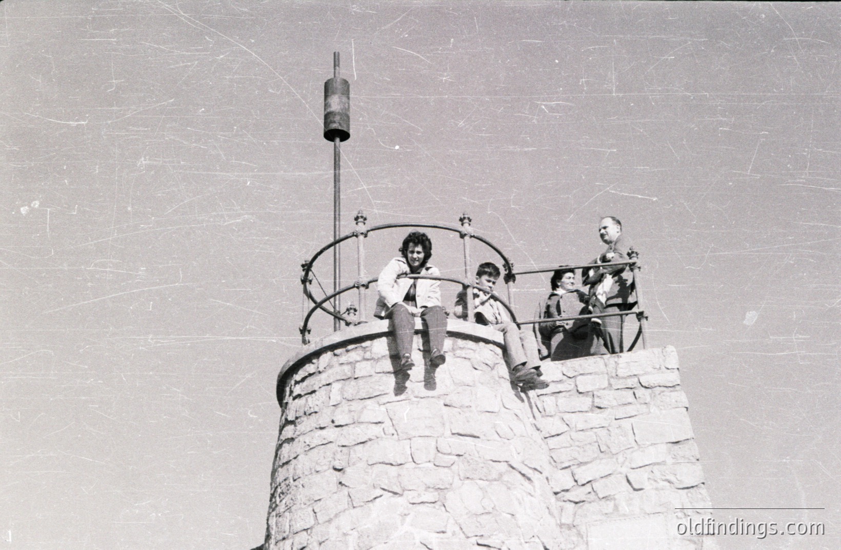A group of four people sit atop a stone observation tower, likely in a mountainous region. The architecture features a circular stone structure with decorative iron railings. Clothing styles suggest the 1960s-1970s. Possibly a tourist location.