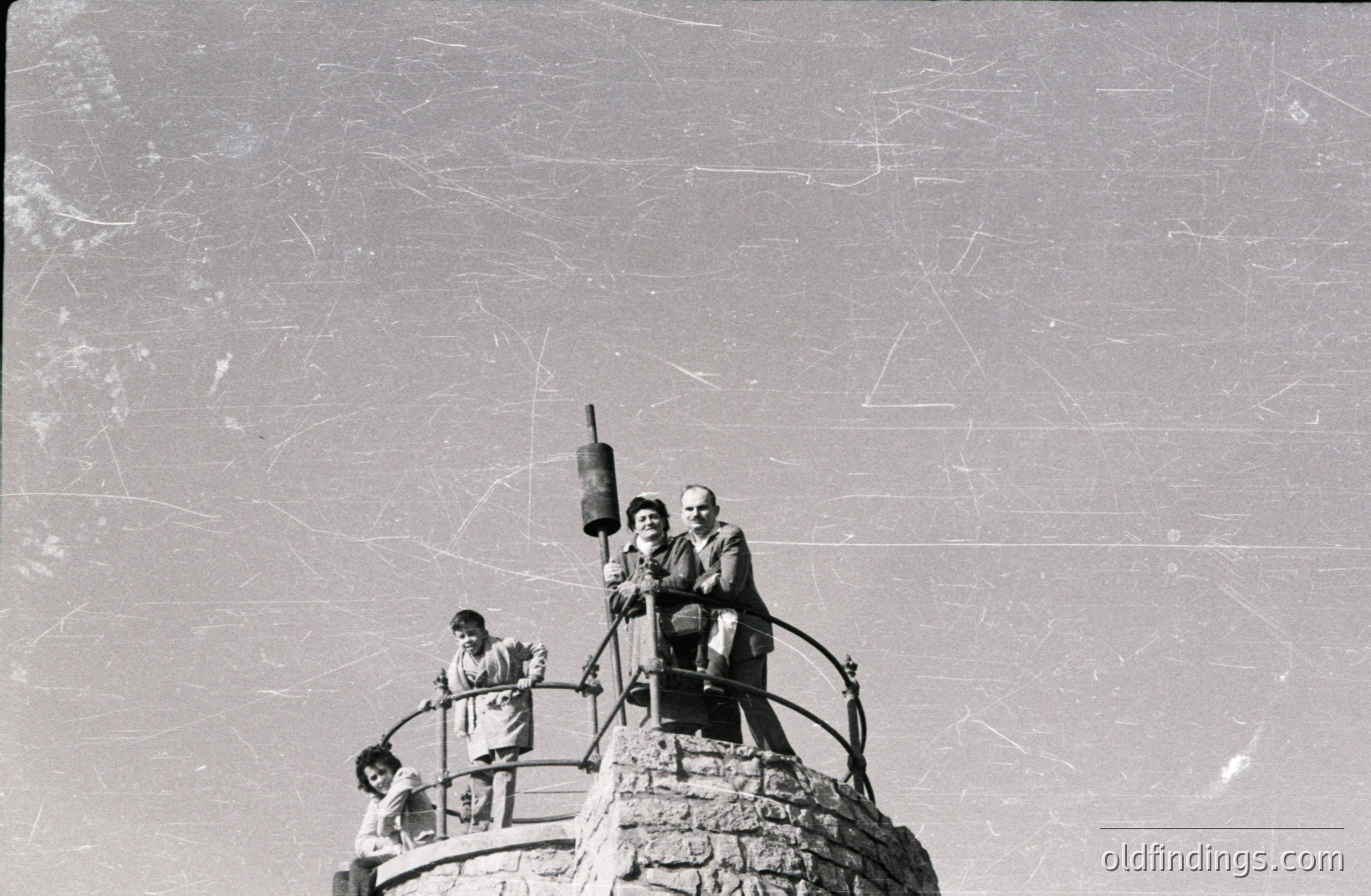 A grayscale photograph depicts three figures atop a stone structure with decorative wrought iron railings. Two adults, a man and a woman, sit on the edge, while a child partially obscures the view from below. The structure appears to be a lookout tower. Likely a tourist attraction. Estimated timeframe: 1940s-1960s.