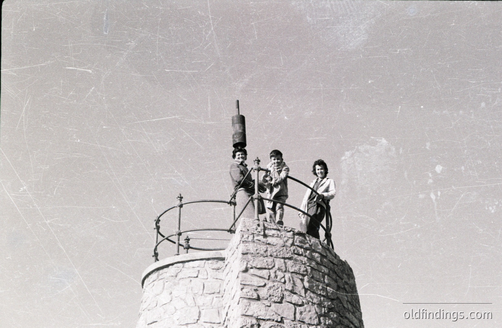 Three children pose atop a stone tower with a metal railing; one playfully balances a bucket on their head. Architecture suggests a European coastal location, likely mid-20th century (1950s-1970s). Likely a souvenir photo capturing youth and leisure.