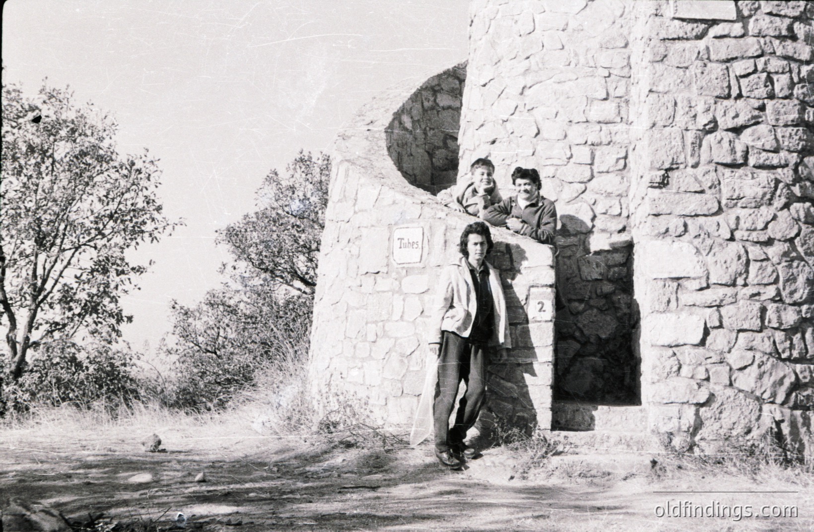 A stone tower anchors the frame, two young men peering from an arched window. The man leaning against the structure wears a corduroy jacket. Sparse vegetation and a dirt path suggest a rural, possibly fortified location. Likely 1970s style. Architectural detail & youthful energy combine.