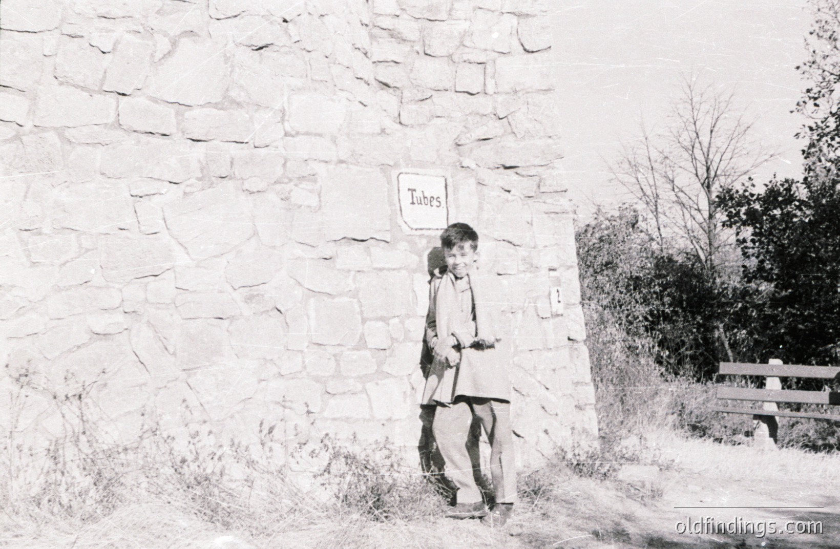 A young boy leans against a stone wall marked "Tubes," with a weathered wooden bench visible in the background. The monochromatic image appears to be a candid snapshot taken outdoors, likely in a rural setting. Appears to be a casual, candid moment. Appx. 1960s-1970s.