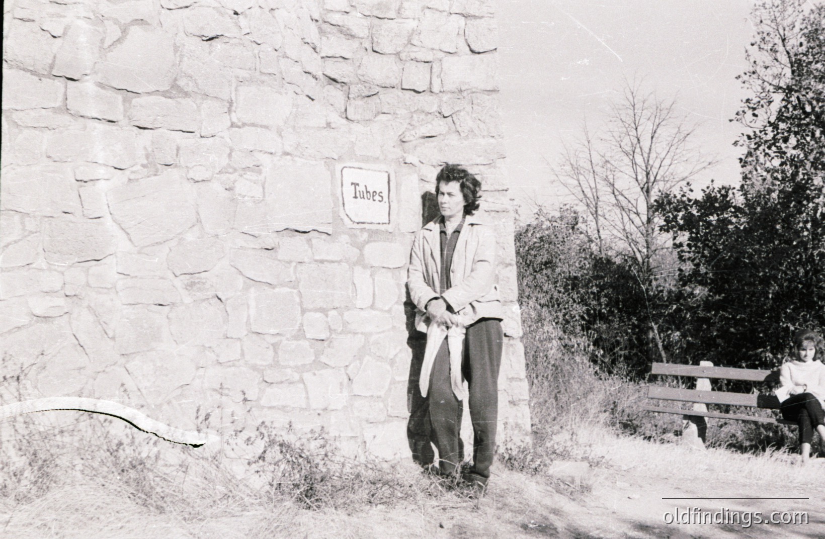 Young man with a layered look stands before a stone wall marked "Tubes." He holds what appear to be bicycle inner tubes. A girl sits on a bench in the background. Appears to be a roadside scene, likely 1960s or 70s. Could be a travel stop or recreational area.