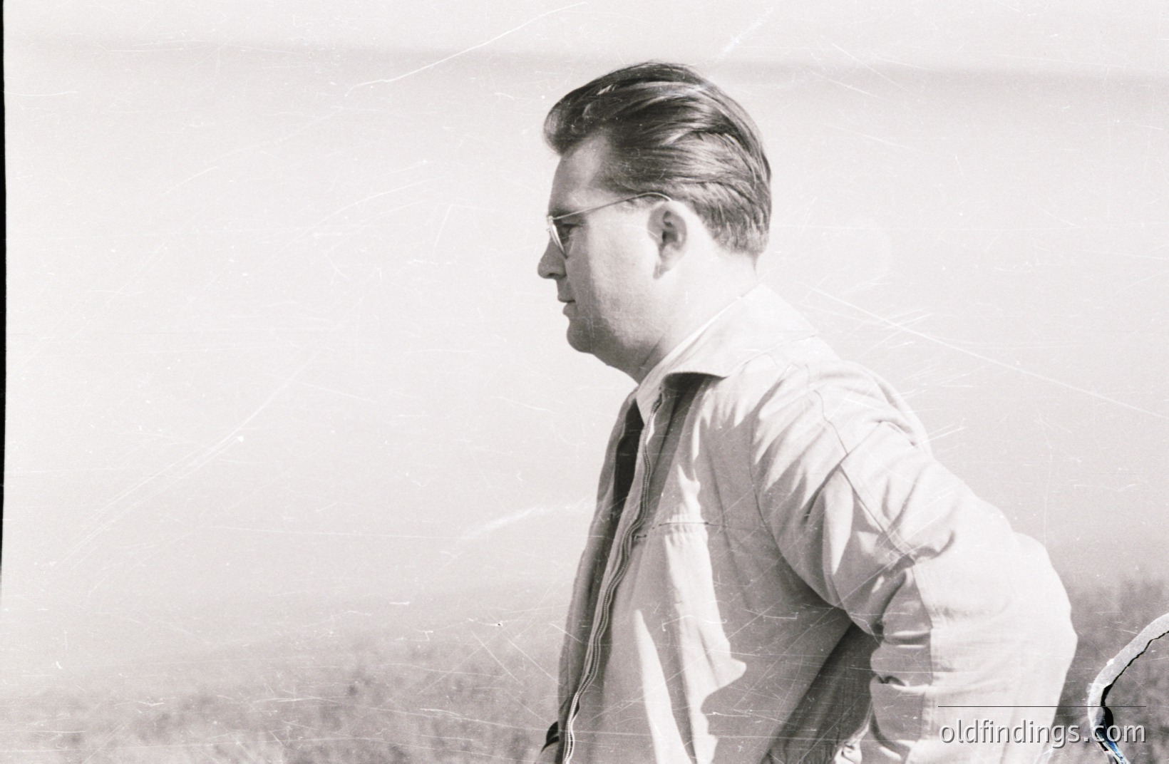 A man in a zipped jacket and tie observes a landscape. He wears glasses and has a neatly styled hairstyle. Appears to be a candid outdoor portrait, likely mid-century, possibly 1950s-1960s. Details suggest a professional or affluent background. Fine grain indicates film photography.
