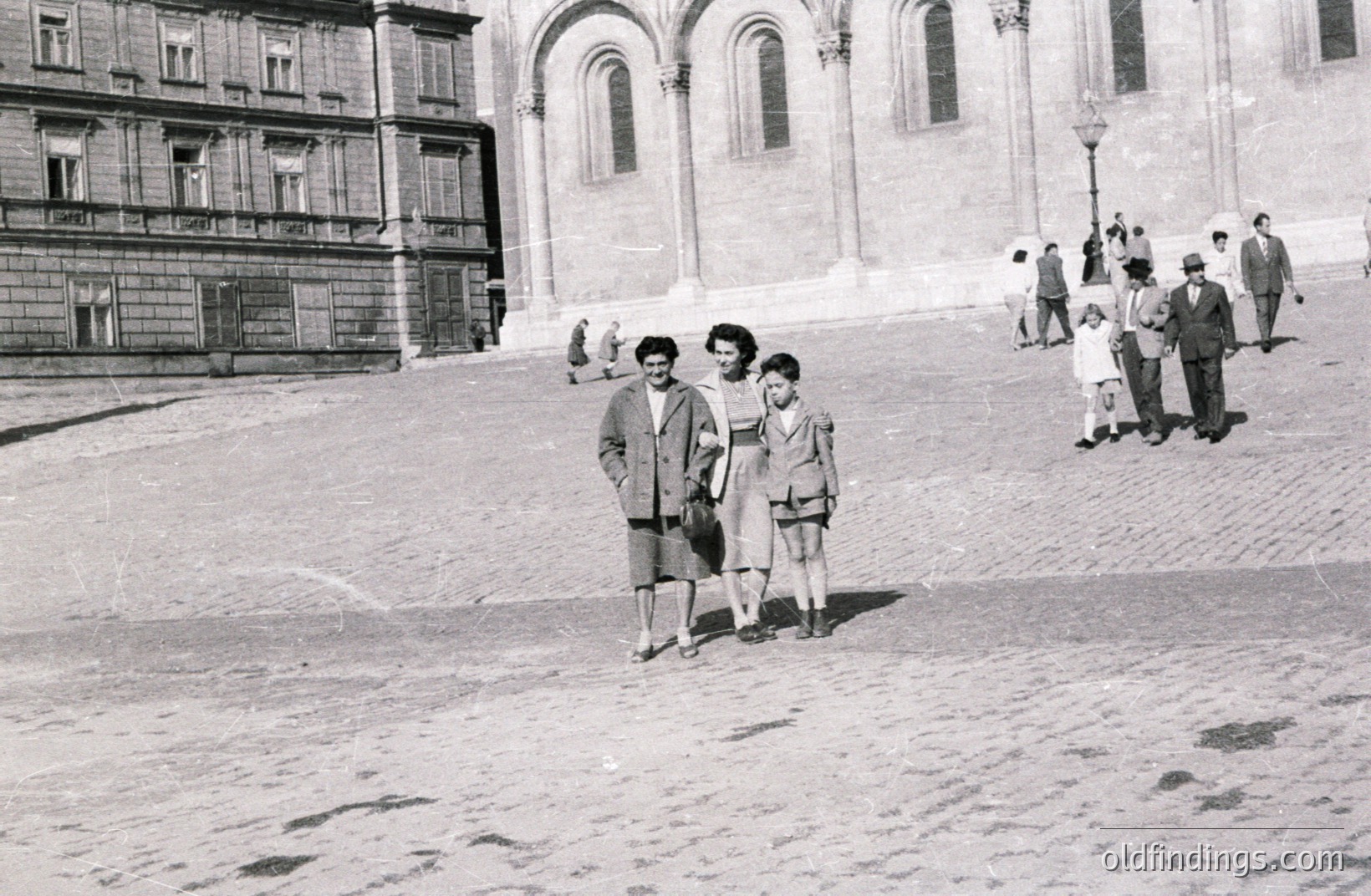 A black and white street scene captures three figures on a wide stone plaza. A woman and a boy, likely related, stand prominently. Several pedestrians are visible in the background, ascending a grand staircase leading to a large building with arched windows. Appears to be a public space. Likely 1950s-1970s.