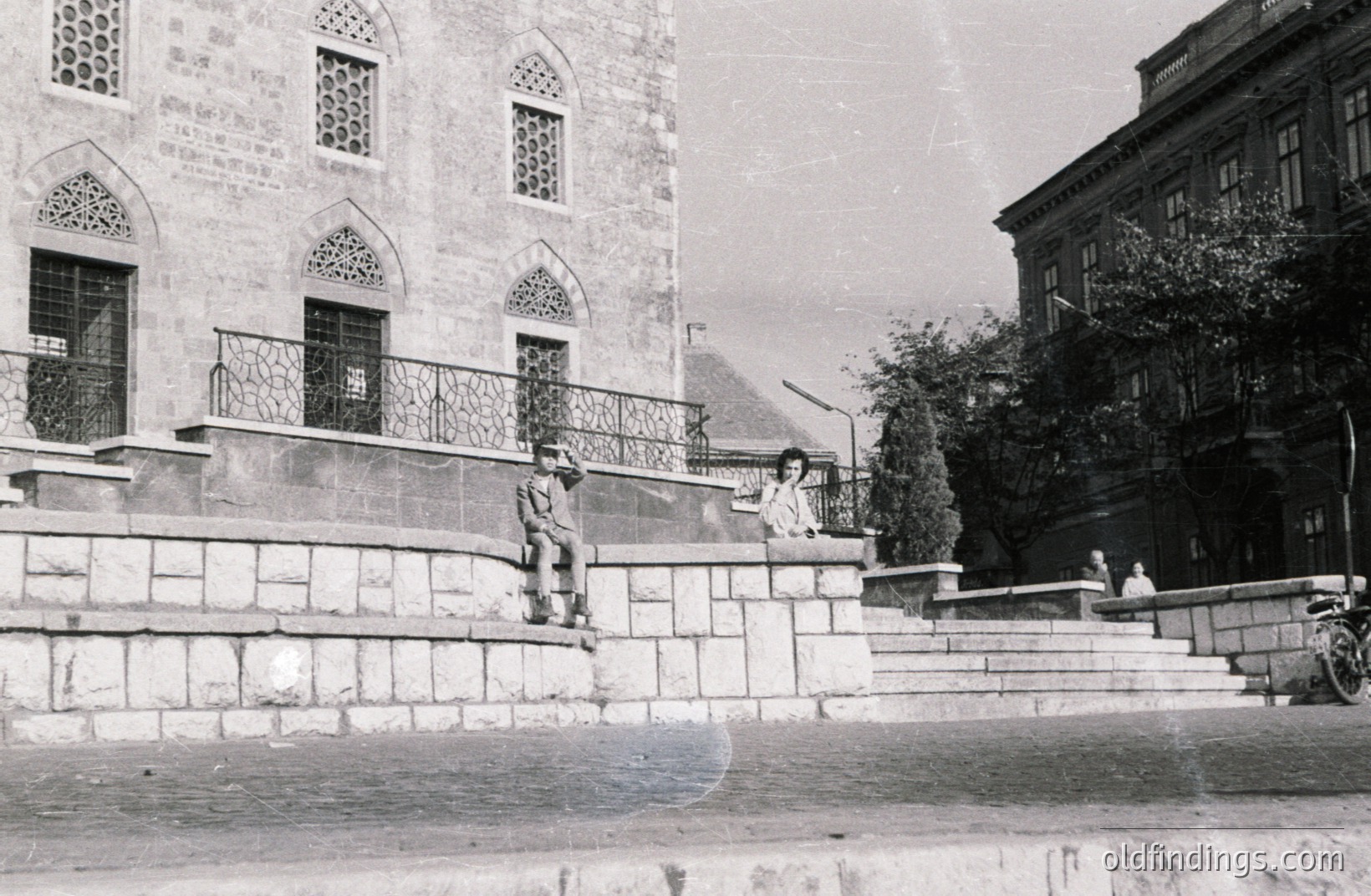 Classic black and white scene; two young people perch on stone steps alongside an ornate building, possibly a public institution or former palace. An older man sits further up the stairs. Likely a European urban setting, perhaps 1960s-70s. Visible vintage scooter and architectural details suggest historical significance.