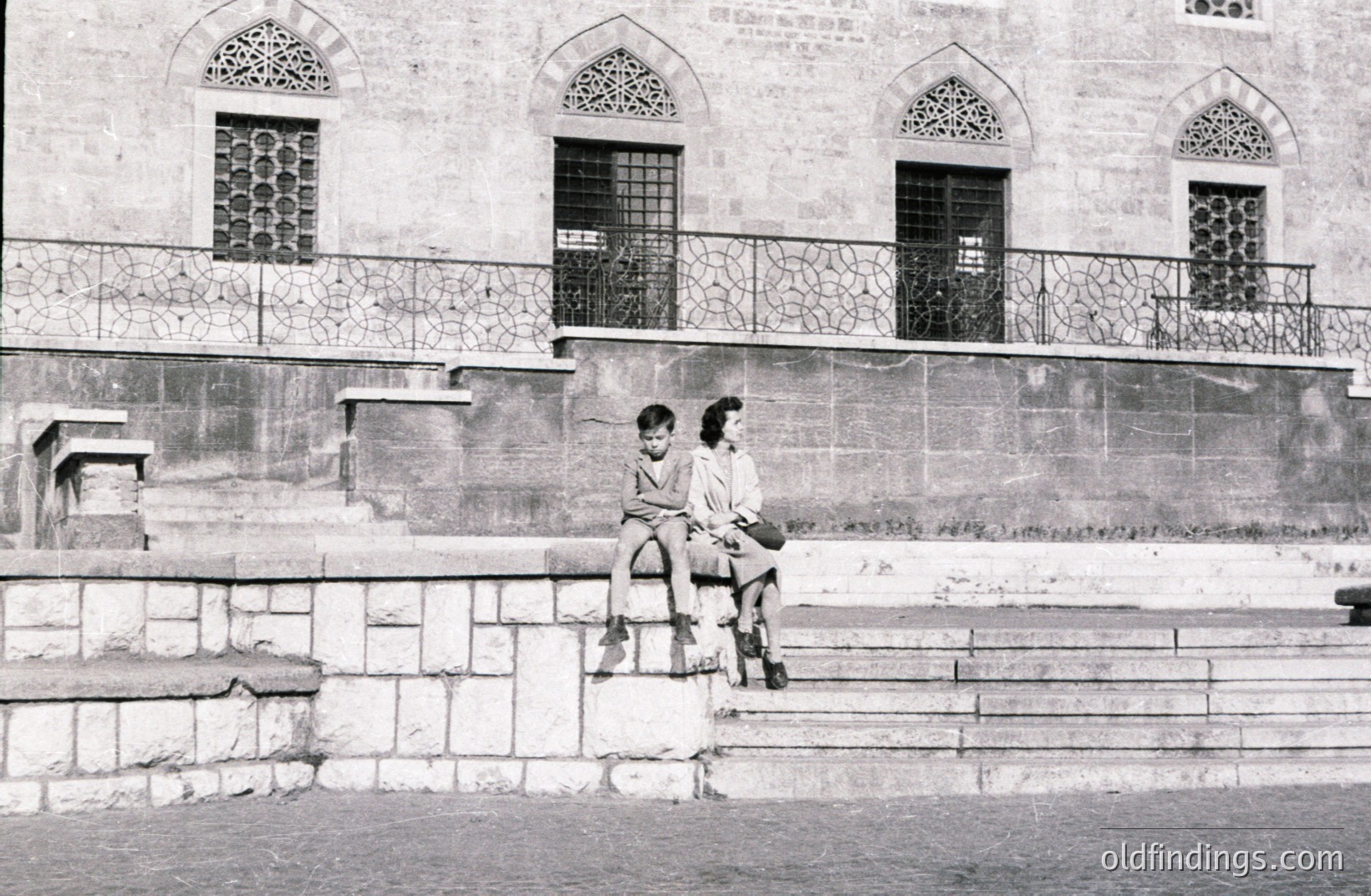 Two children sit perched on a stone staircase leading up to a building with ornate arched windows and a decorative metal balcony. The structure appears to be of historical significance, possibly a public building or residence. Likely 1950s-1970s, location uncertain. Stone architecture and clothing styles are defining features.