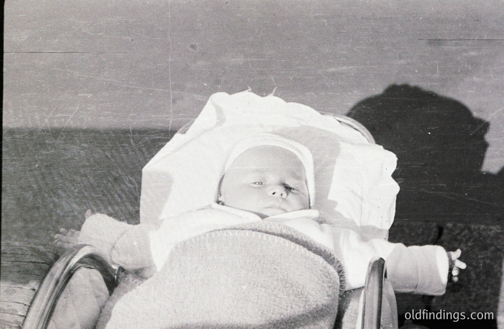 Infant asleep in a vintage pram, bundled in blankets & a close-fitting bonnet. A shadow is cast from the pram's frame. Likely a family snapshot, circa 1930s-1950s. Evokes nostalgia and early childhood imagery. Excellent potential for design or historical contexts.