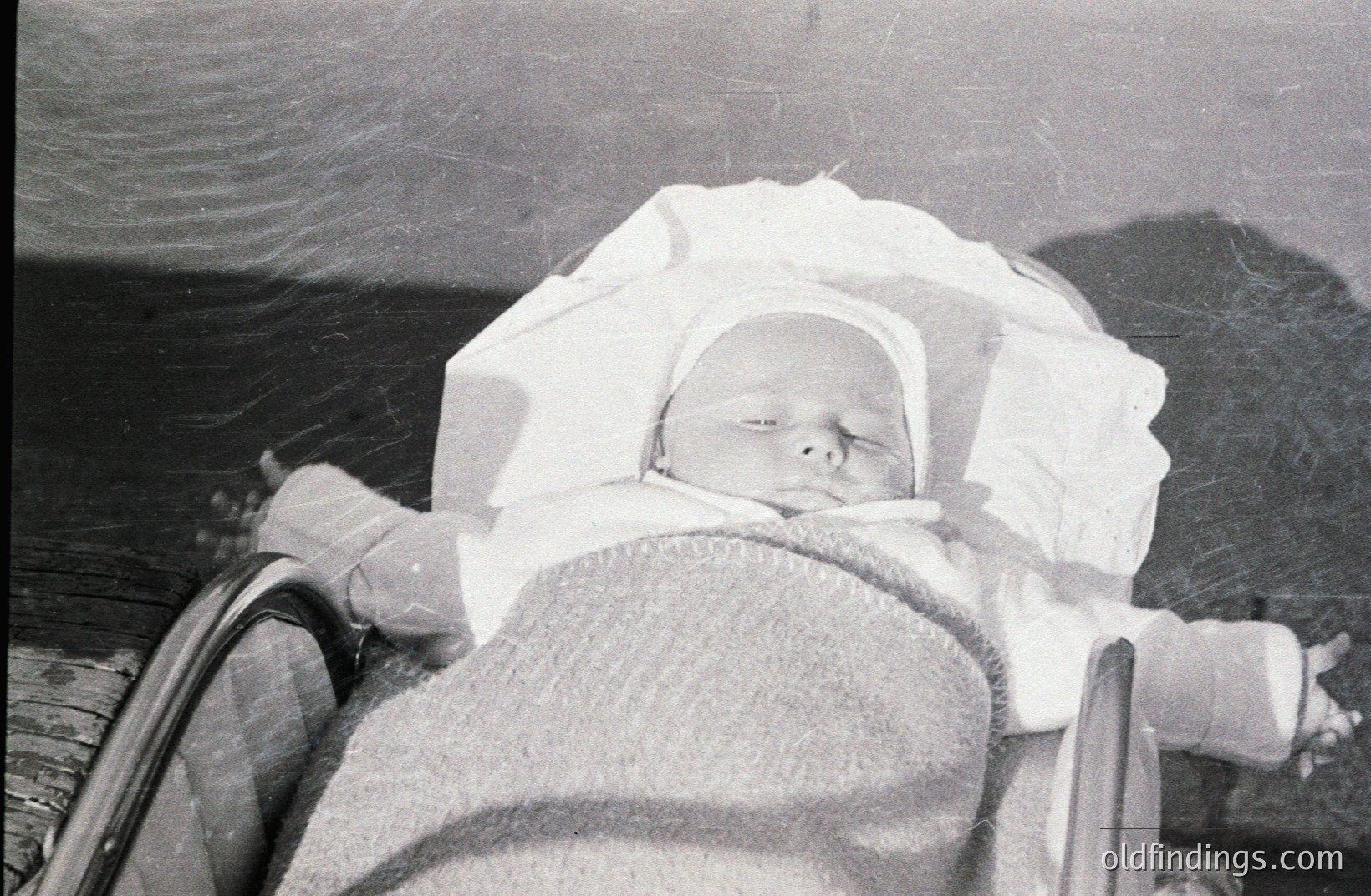 Infant sleeping peacefully in a vintage wicker cradle, swaddled in white cloth and a bonnet. Likely a family snapshot from the mid-20th century, possibly 1940s-1960s. The cradle’s construction suggests a period of handcrafted nursery furniture. Shows a moment of quiet domesticity.