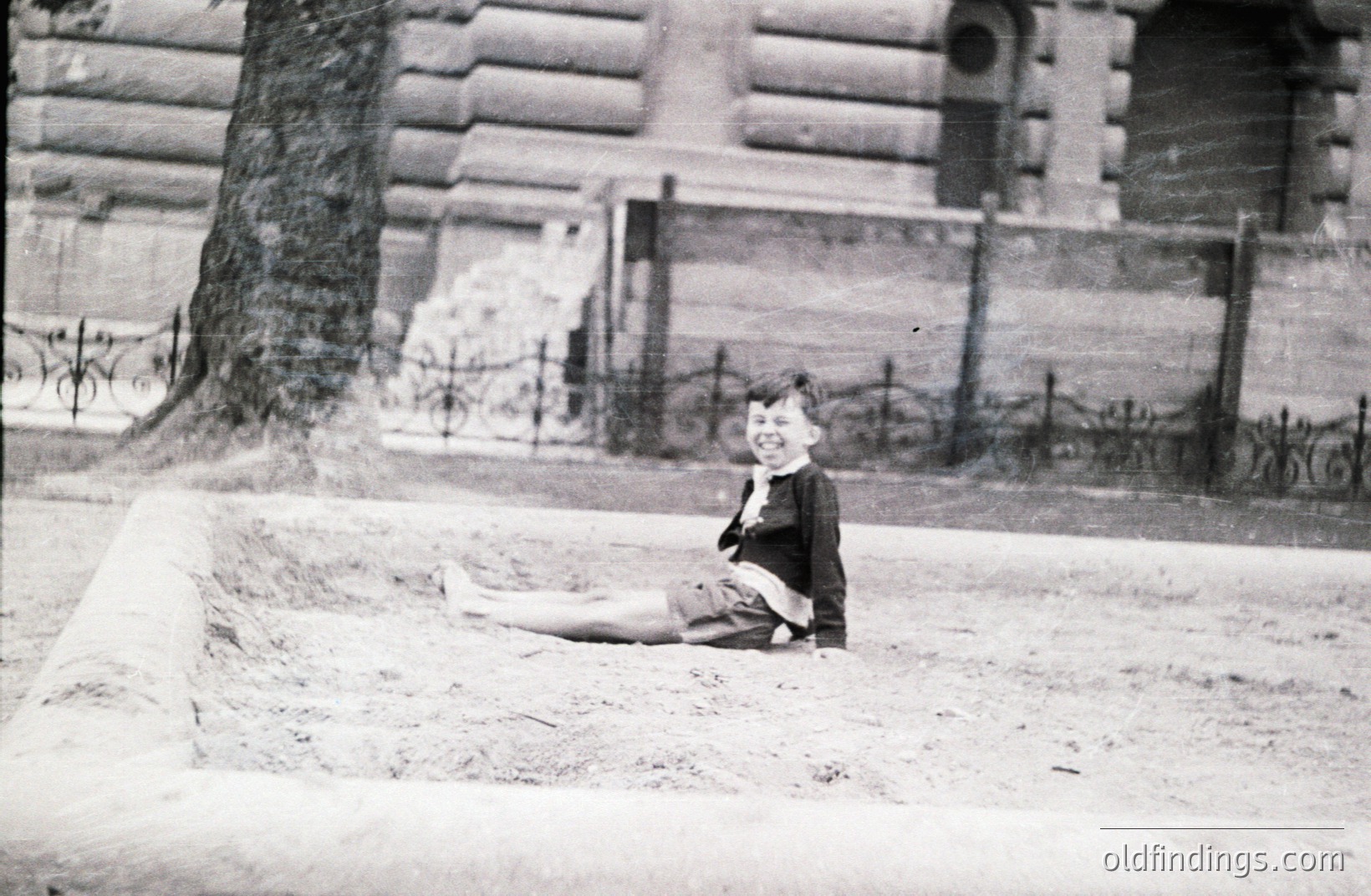 A young boy sits in a shallow depression on sandy ground, wearing a dark jacket and shorts. Ornate wrought-iron fencing and a grand building with arched windows form the backdrop. Likely taken in the early 20th century, possibly a formal garden setting. A charming glimpse of childhood.
