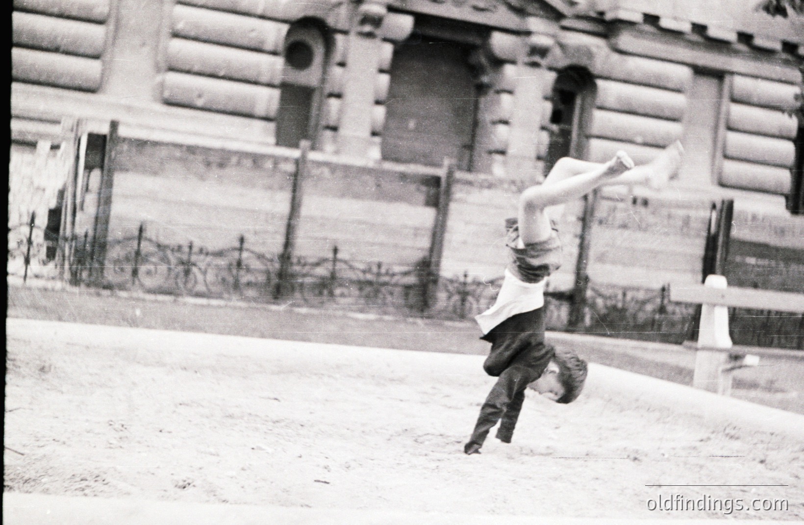 A young boy performs a handstand on a slightly sloped, gravel surface. He wears short pants and a button-down shirt. A detailed stone building with arched windows forms a backdrop. The image exhibits vintage photographic qualities, suggesting a mid-20th century timeframe. Likely a candid street scene.