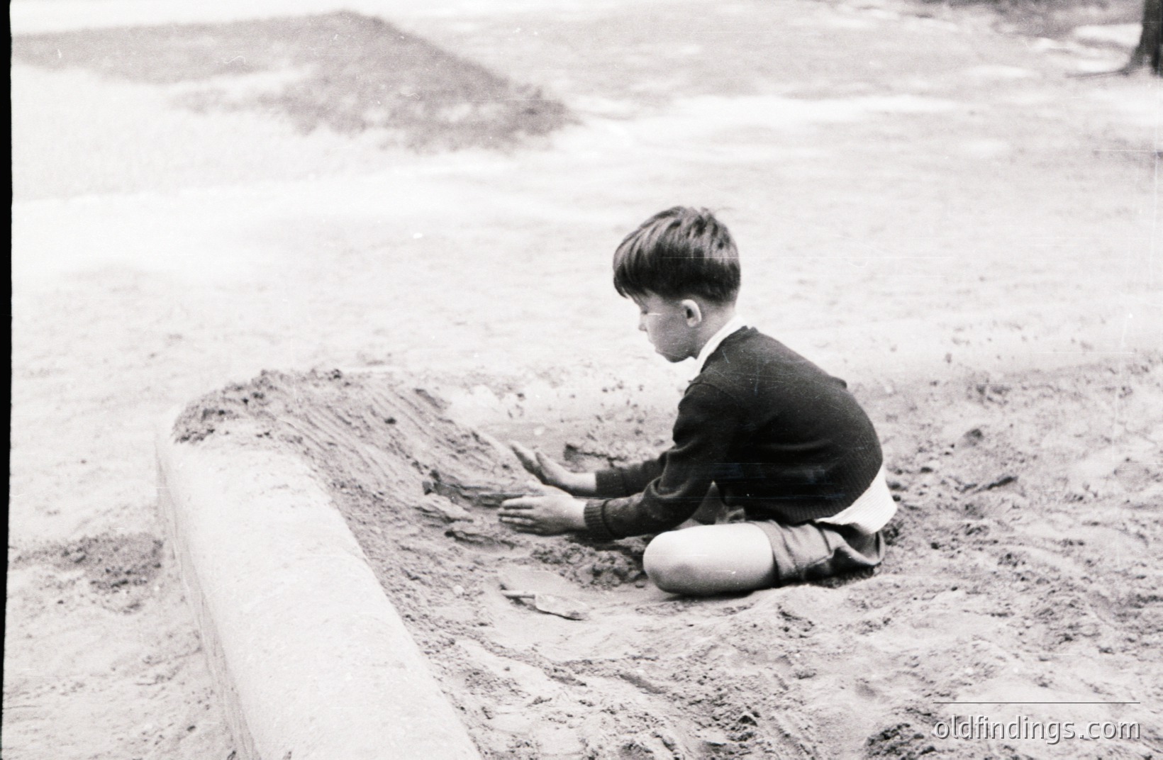 A young boy, dressed in a collared shirt and sweater, kneels within a shallow, concrete-lined depression, possibly a drainage ditch. He’s engaged with the muddy earth. Appears to be a candid moment, circa 1950s-1970s. Potential stock value for childhood, play, or urban landscapes.