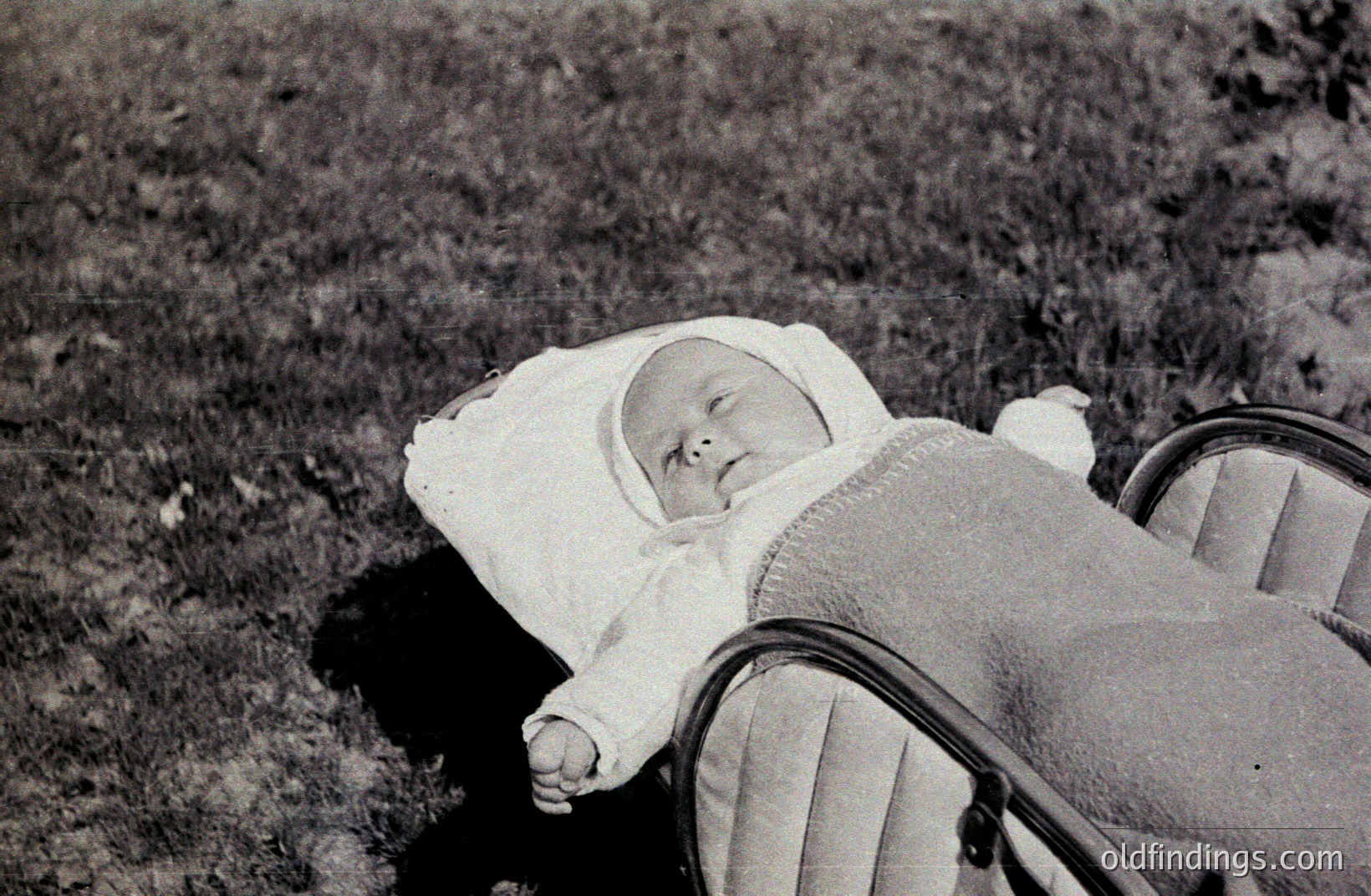 A baby, bundled in a white bonnet and blanket, rests in a vintage pram on a grassy lawn. The pram’s frame is visible, showcasing a cushioned seat. Likely a family snapshot from the mid-20th century (1950s-1970s), evokes a nostalgic, domestic scene. Good for heritage projects.