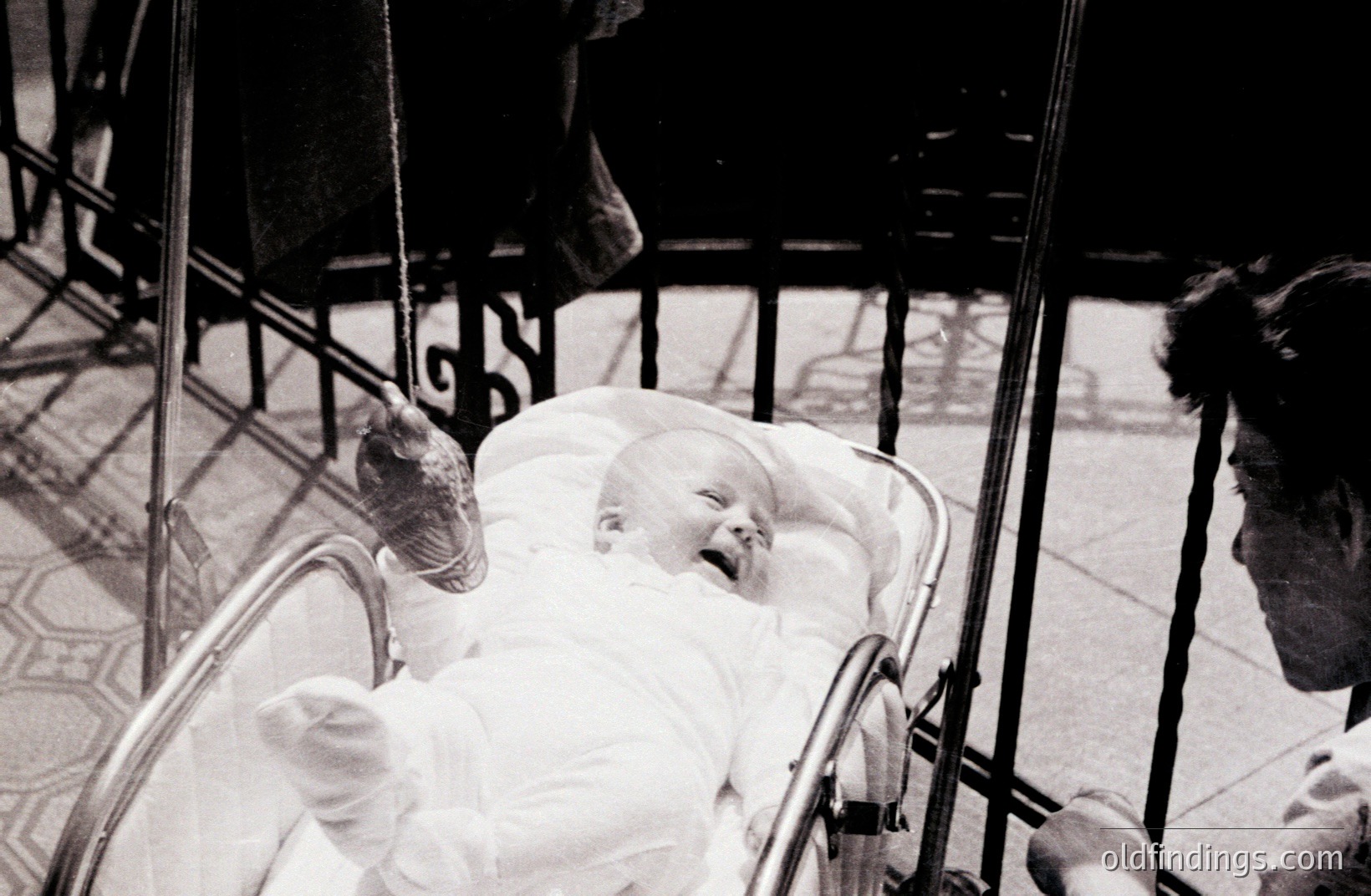 Infant in a stroller, likely outdoors, with patterned tile flooring visible. The child, dressed in white, appears to be laughing or crying. Adult’s hand and partial view of attire are seen near the stroller. Simple composition, possibly 1960s. High potential for design use.