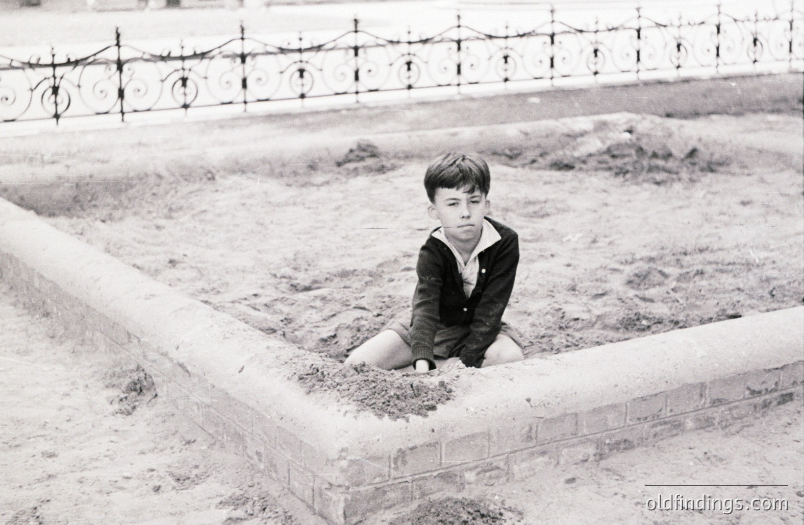A young boy, seated within a newly constructed brick-lined depression or garden bed. He wears a collared shirt and dark blazer. A decorative, wrought-iron fence forms the backdrop. The image’s starkness suggests a mid-20th century, potentially 1950s, documentary or snapshot. Potential design reference for childhood imagery.