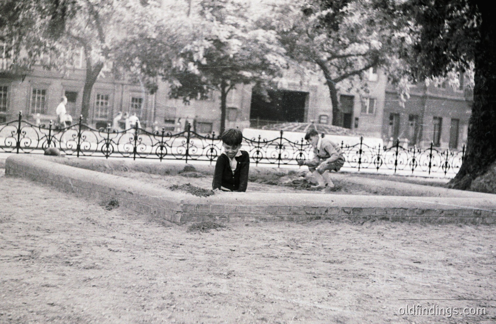 Two boys engaged in play within a walled park area. One kneels, examining the ground; the other sits, using a tool to manipulate the soil. Background features a formal building facade and ornate wrought-iron fencing. Likely mid-20th century, documenting childhood recreation.