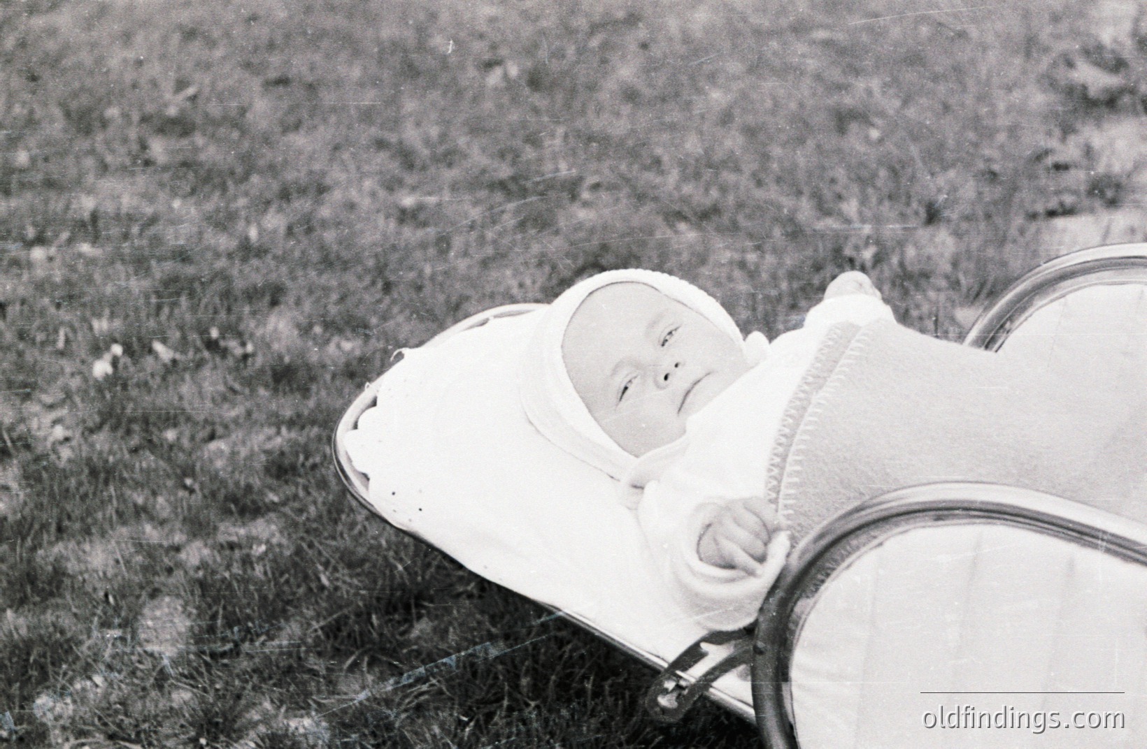 A baby, swaddled in white cloth and wearing a bonnet, reclines peacefully in a vintage stroller amidst tall grass. The image, captured in black and white, suggests a candid moment. Likely from the mid-20th century, this photo holds sentimental value and could be used for nostalgic design or historical family research.