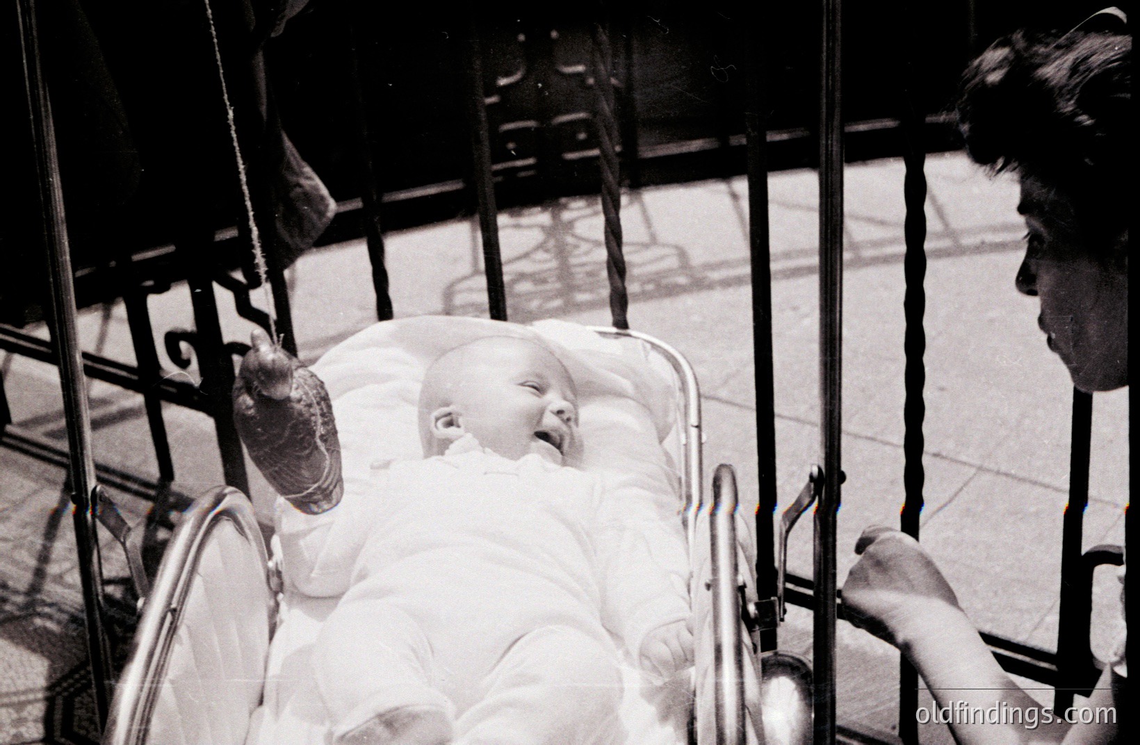 A baby, dressed in white, rests in an ornate, vintage pram. A blurred figure, presumably the caregiver, is partially visible in the background. Black and white film suggests a mid-20th century era. Likely a candid street portrait; evokes a sense of domesticity & fleeting moments.