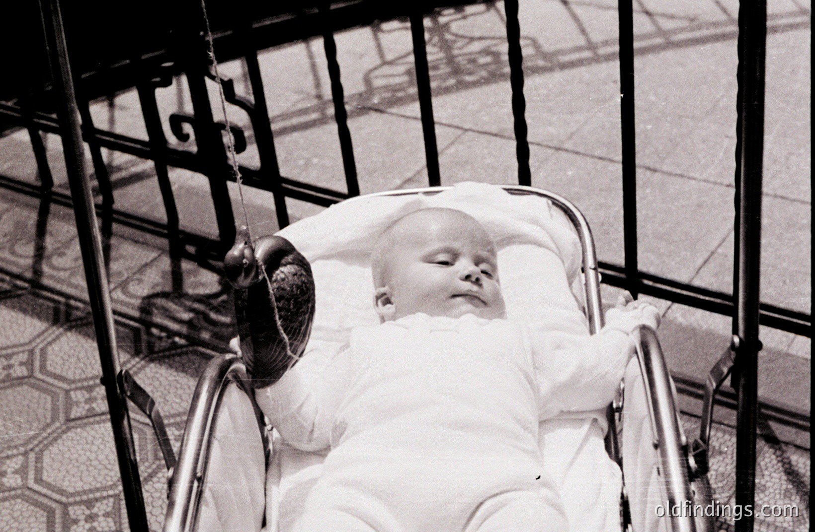 A serene infant, swaddled in white, sleeps peacefully within a vintage chrome and rattan pram. The stroller sits before a wrought-iron gate and patterned tiled patio. Likely a domestic scene, possibly 1950s-1970s. Evokes nostalgia and childhood innocence. Could be valuable for design or vintage advertising.