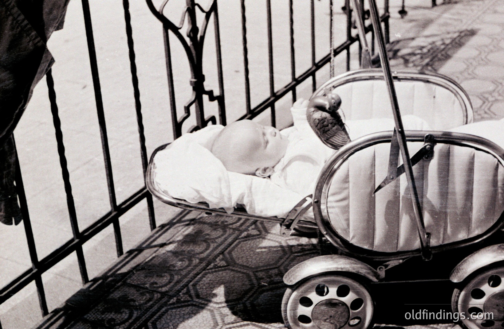 Infant peacefully rests within an ornate, vintage baby carriage, set against a wrought-iron railing. The carriage's design showcases circular wheel hubs and a detailed frame. Patterned floor tiles suggest a courtyard or terrace setting. Likely 1950s-1970s, possible European origin.