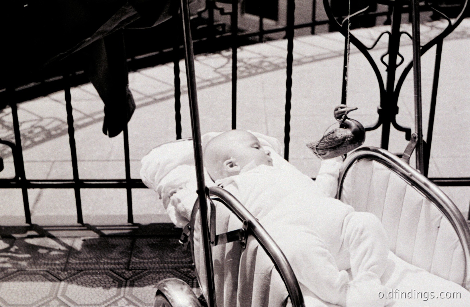 A baby sleeps peacefully in a vintage stroller, perched within wrought iron railings. A pigeon has landed on the stroller's hood, seemingly undisturbed by the infant. Likely 1960s or 70s era, hinting at stock photo or design reference potential. Evokes a quiet, domestic moment.