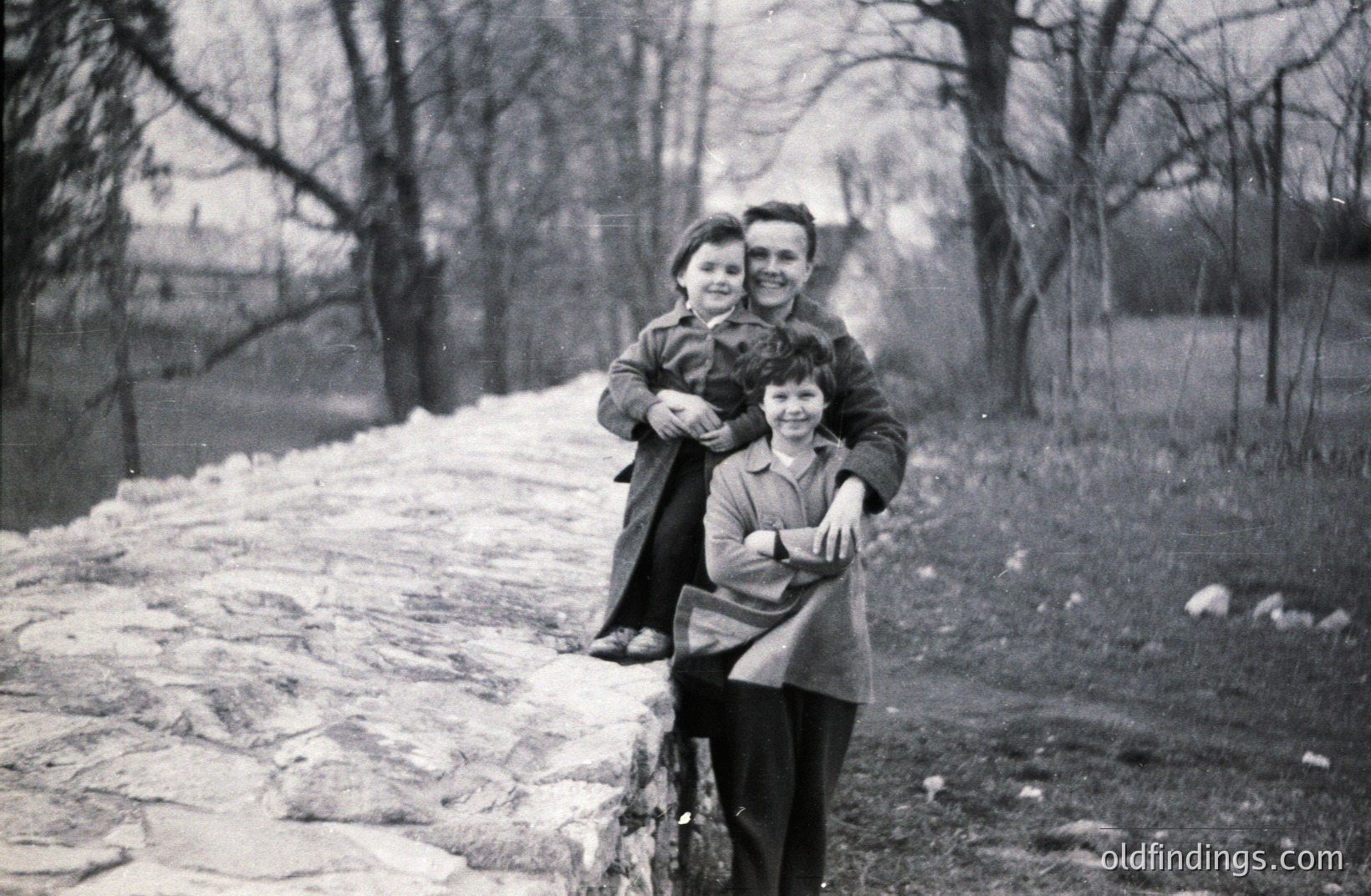 A young boy sits on a man’s shoulders atop a stone retaining wall, set against a backdrop of bare trees. Both are wearing mid-century outerwear. Likely 1950s or 60s, possibly a rural setting. Image has documentary, family portrait value.