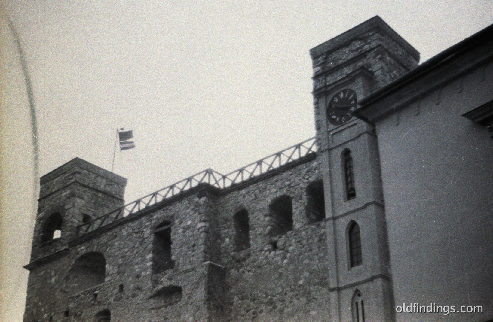 Monochrome photograph depicts a stone fortress or castle wall with a clock tower, likely European. Open crenellations line the top of the wall, and a flag waves. Possible seaside location. Architectural style suggests late 19th or early 20th century. A valuable reference for design or historical research.