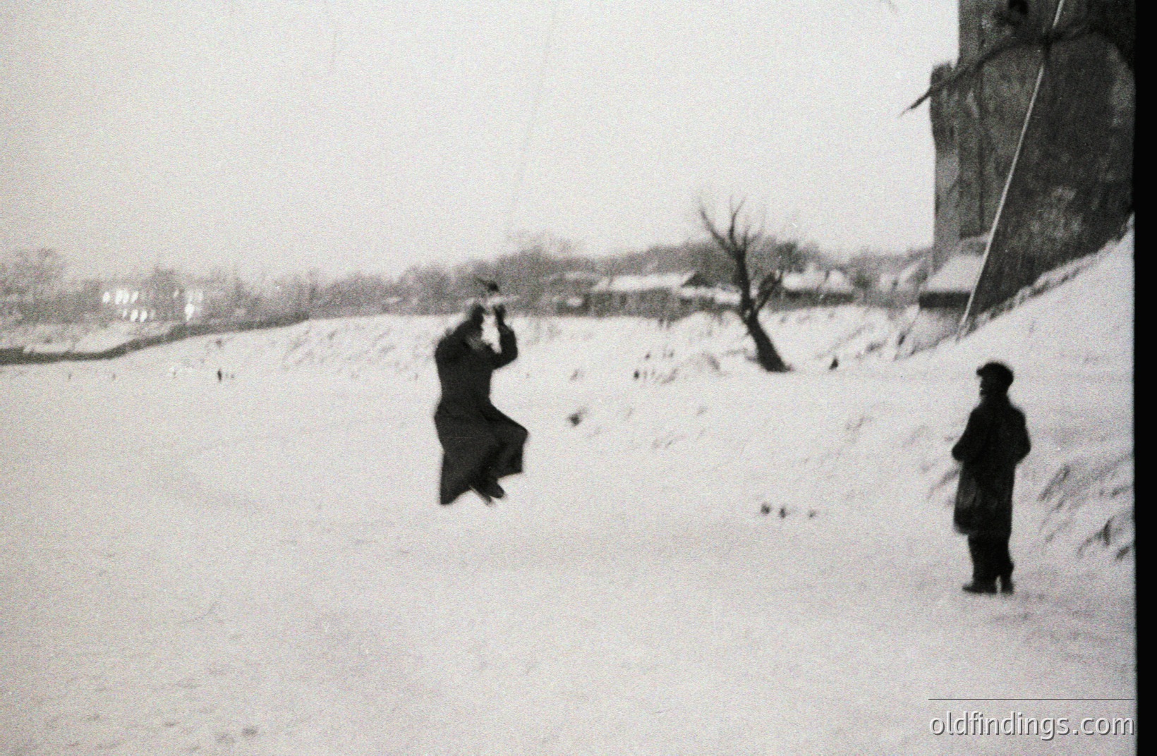 A man in a dark coat and hat leaps into a snowy landscape, seemingly propelled upwards. A second man stands observing, slightly distanced. A hillside rises in the background, with visible buildings and sparse vegetation. Appears to be an early photographic experiment, potentially documenting movement.