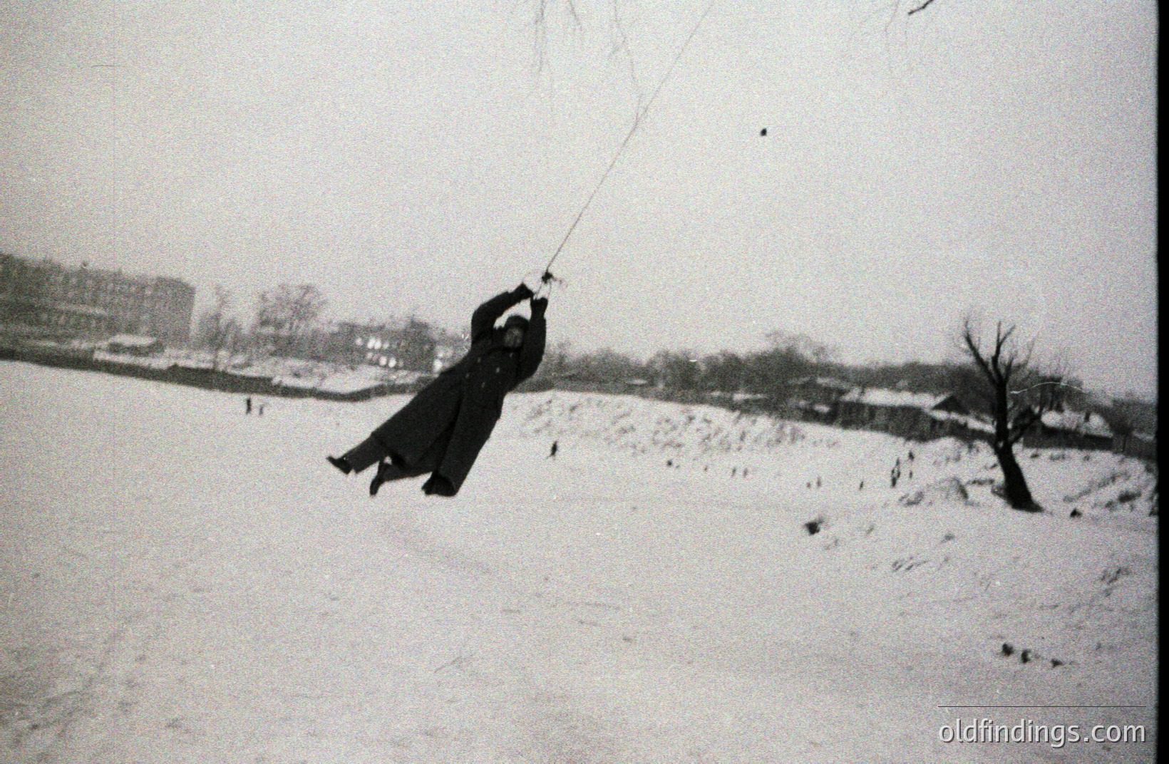 A person in a long coat swings from a rope across a snow-covered field. The pose suggests mid-swing, with blurred motion. Background shows buildings and bare trees under a wintry sky. Likely early 20th century documentation of leisure activity.