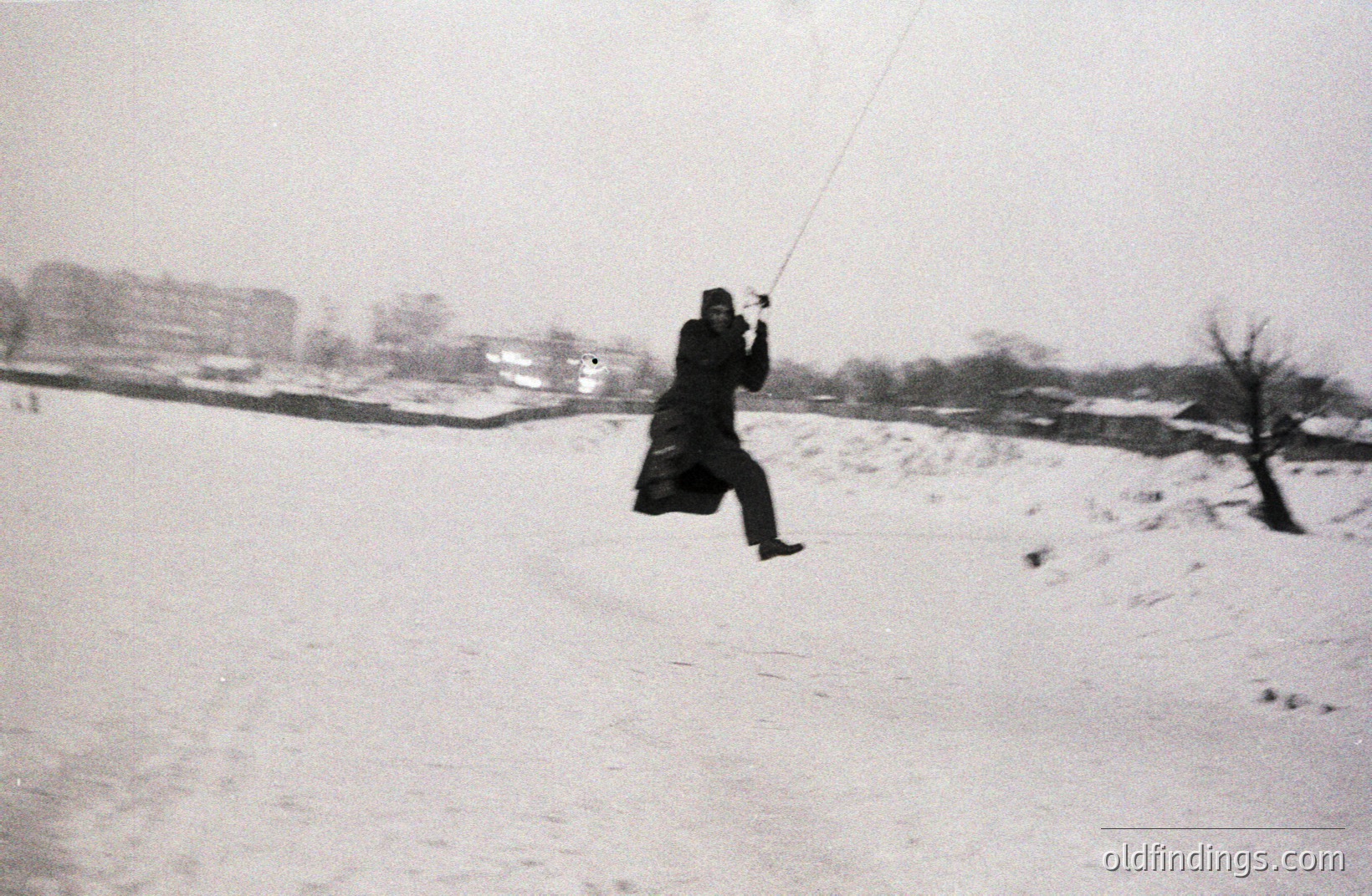 A person, bundled in a dark coat and hat, leaps mid-air while suspended from a rope. Snowy landscape with apartment buildings visible in the background. Appears to be a candid moment, possibly a playground or open field. Likely 1970s or 1980s style clothing.