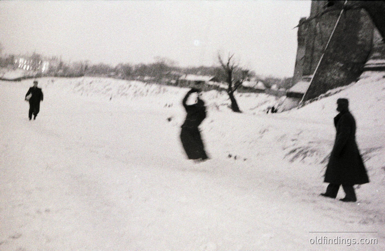 A monochrome street scene captures three figures amidst a snow-covered landscape. Two individuals, cloaked in dark coats, are visible – one walking away, the other in mid-motion, arms raised. A stark, angled stone structure rises on the right. Likely taken in the mid-20th century. Potential stock photo value for design or historical context.