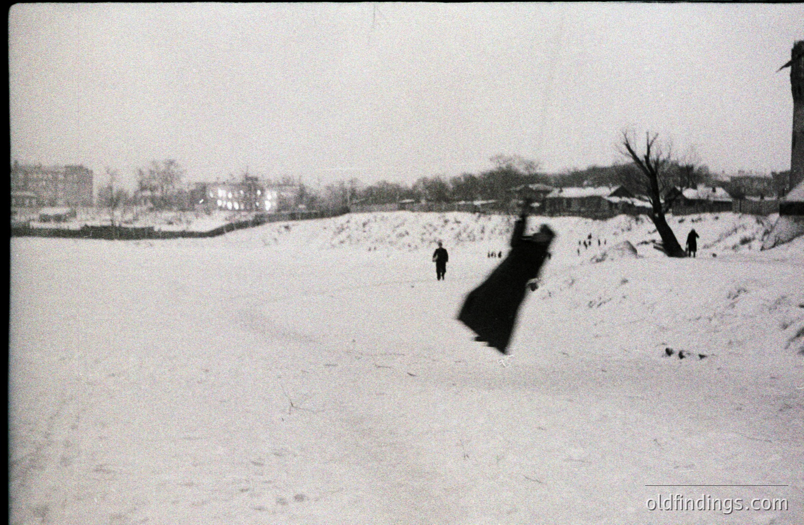 Snow-covered landscape features a slight rise in the ground. Three figures are sparsely placed across the scene. A single, dark coat is lifted high by the wind. Architectural details suggest a mid-20th century European town. The image exhibits visual texture and stark contrast.