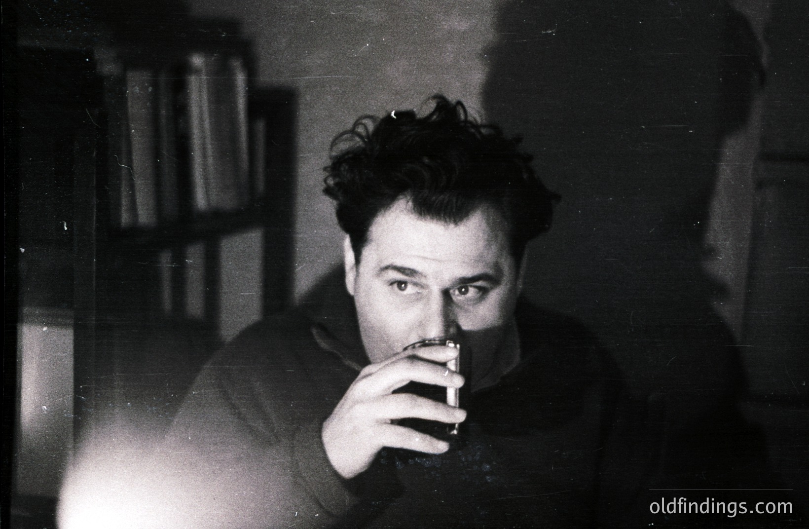 Intimate portrait of a man with tousled hair, holding a drink and lit cigarette. Bookshelves fill the blurred background. Stark lighting emphasizes the subject's gaze and adds a moody, contemplative atmosphere. Likely a candid, 1960s-era photograph.