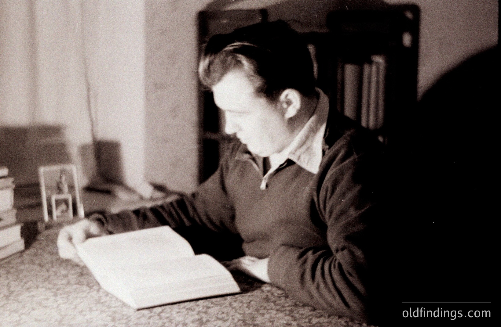 A young man, seen in profile, intensely reads a large, open book seated at a table. Shelves of books form a blurred backdrop. He wears a collared shirt and a dark sweater. Likely a student or scholar, capturing an academic moment. Estimated 1950s-1970s.