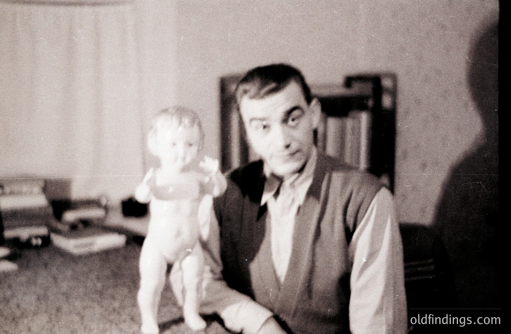 A man in a button-down shirt & vest poses with a small, plaster statuette of a child. The setting appears to be a room with bookshelves. Appears to be a snapshot, potentially 1950s-1970s. A somewhat formal, yet intimate portrait.