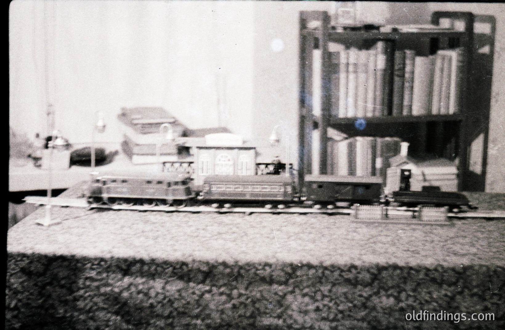 Detailed black and white photograph shows a miniature train set displayed on a table, with a large bookcase in the blurred background. Includes a locomotive, passenger car, and a small freight wagon. Likely a hobbyist’s display, circa 1950s-1970s.