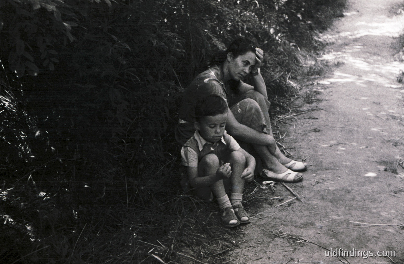 A mother and young boy sit nestled against foliage on a dirt path. The boy wears sandals and a collared shirt, while the mother’s face shows weariness. Likely captured in a rural setting, this poignant image conveys resilience and hardship. Appears to be 1950s-1970s. Valuable for social documentary and historical research.