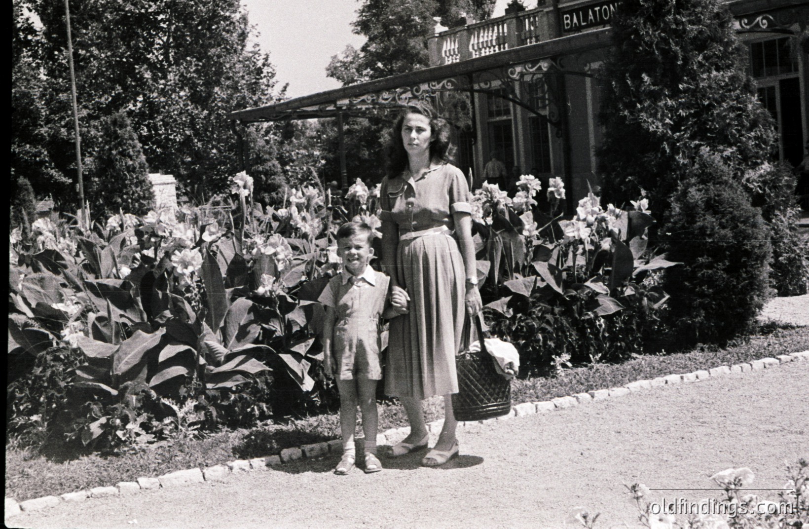 Woman and young boy pose near a grand villa, likely lakeside. The woman carries a picnic basket, showcasing mid-century fashion. The architecture suggests a European resort setting. Estimated period: 1950s-1960s. Possibly a vacation snapshot or stock photo reference.