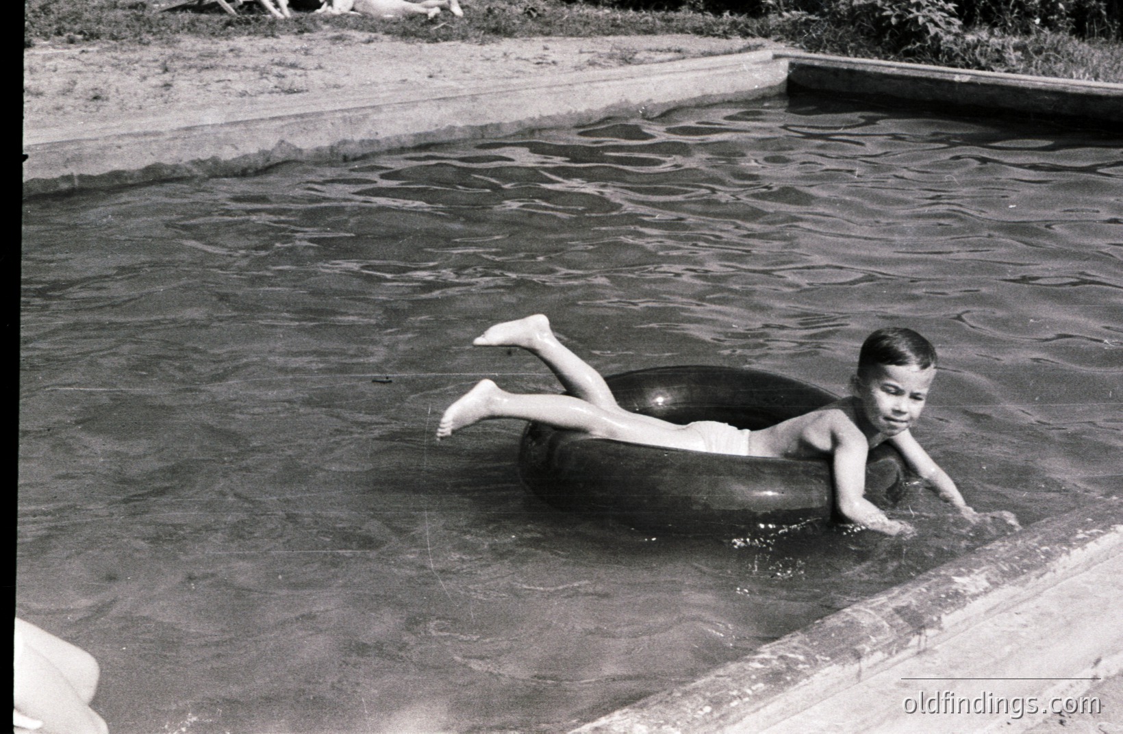 A young boy floats on an inflatable inner tube in a swimming pool. He appears to be paddling with his hands while submerged. Concrete edging surrounds the pool; figures are blurred in the background. Likely a family vacation snapshot, circa 1960s-1970s Americana. Stock potential for nostalgic summer themes.