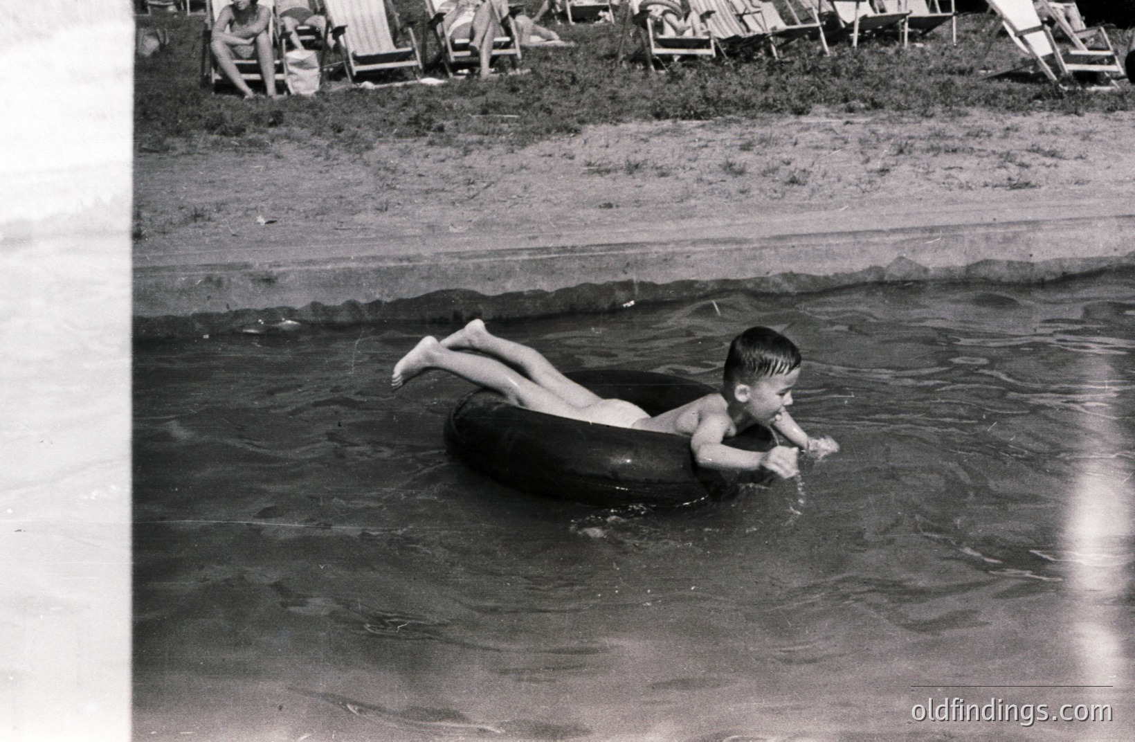 A young boy floats on a black inner tube in calm water. Background shows a beach with multiple lounge chairs. Appears to be a candid snapshot, likely from the 1960s or 70s. Simple, nostalgic depiction of summertime leisure. Possible use for stock imagery or vintage design.