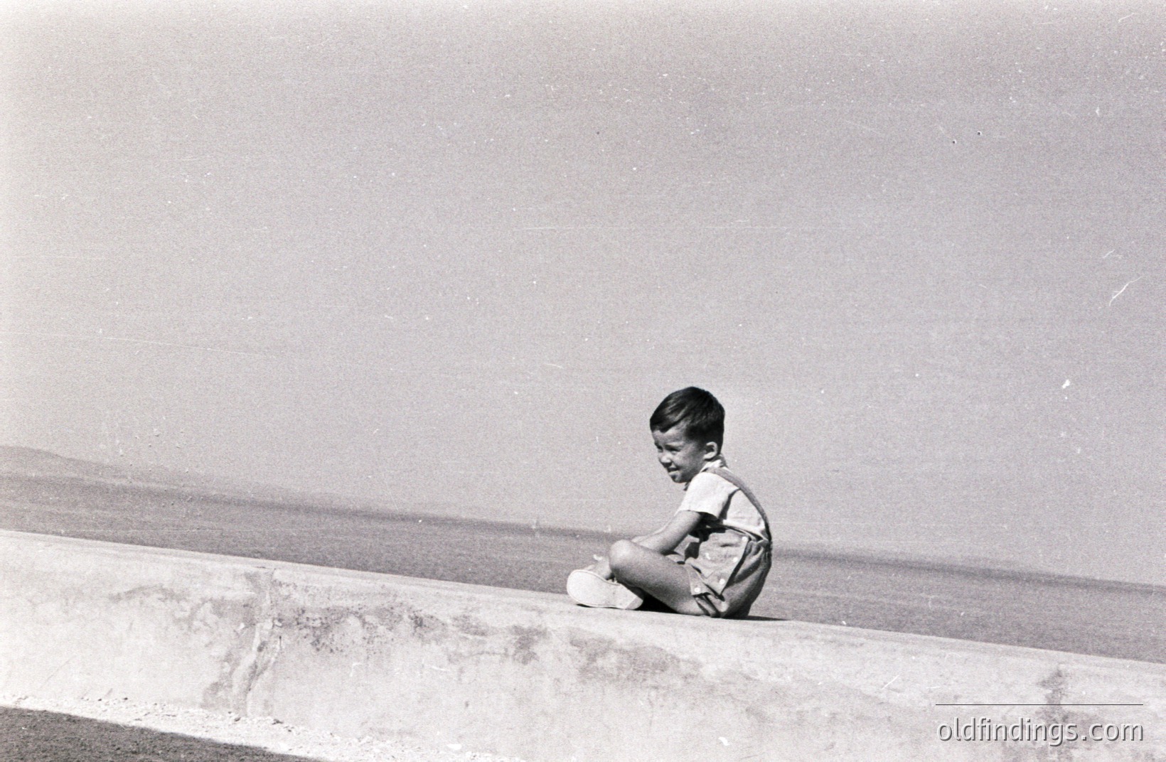 A young boy sits on a concrete retaining wall overlooking the sea. He wears short shorts and a short-sleeved shirt, appearing contemplative. The scene suggests a seaside location, likely a coastal town or resort. The photograph's style & grainy quality suggests the 1960s or 1970s. A candid portrait of childhood and leisure.