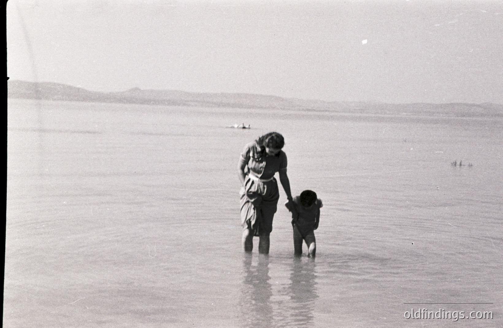 A woman and young child are seen wading in shallow water, possibly a sea or lake. The scene appears calm, with distant hills visible on the horizon. Clothing style suggests a mid-20th century timeframe. Likely a candid moment of family recreation.